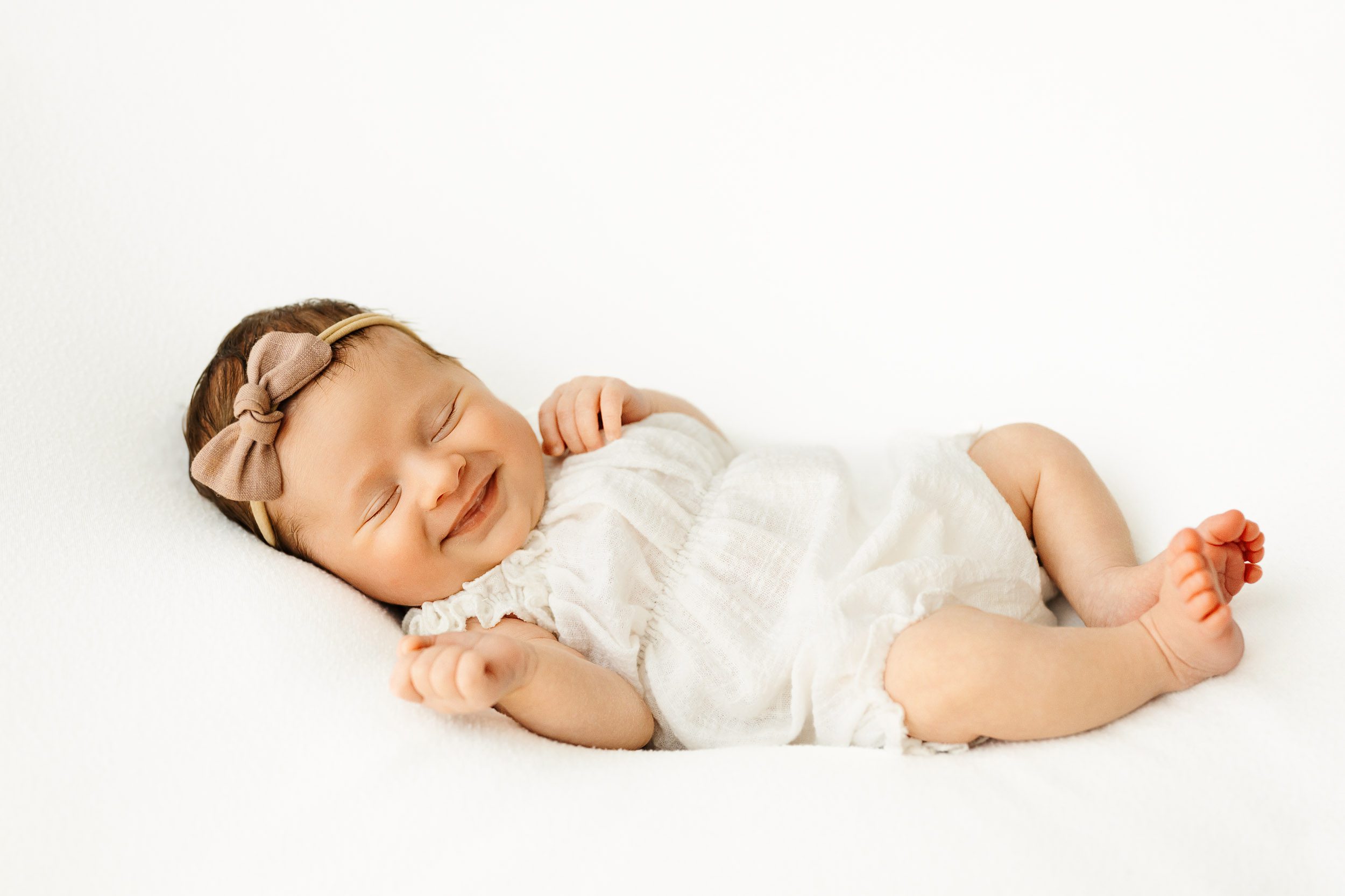 a baby girl wearing a white linen romper laying on a white backdrop with a sleepy smile on her face during a natural newborn photography session