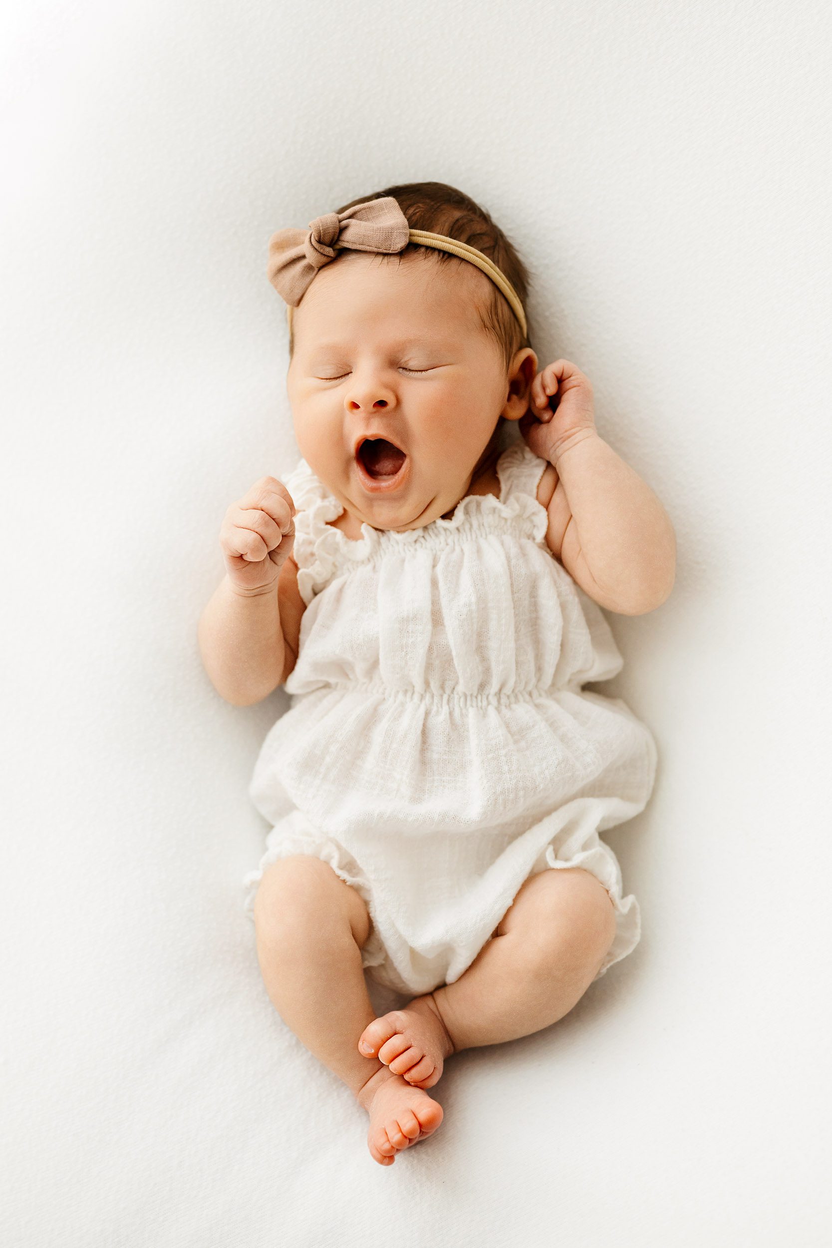 a baby girl wearing a white linen romper laying on a white backdrop and yawning during a natural newborn photography session