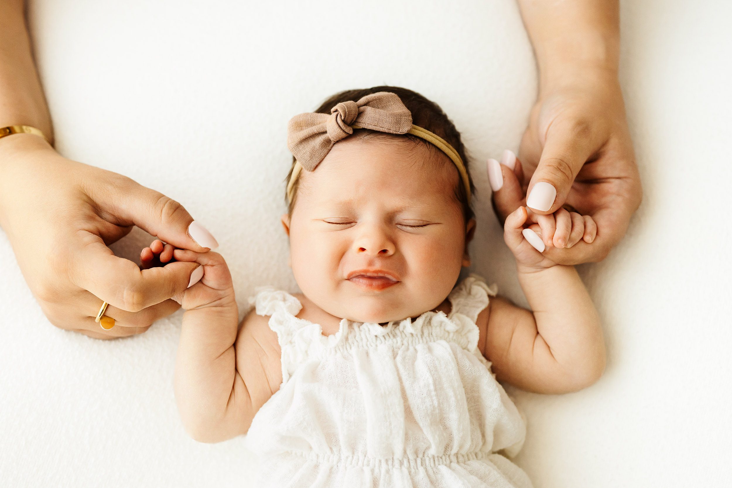 a baby girl wearing a white linen romper laying and holding onto her mom's fingers with her tiny hands during a natural newborn photography session