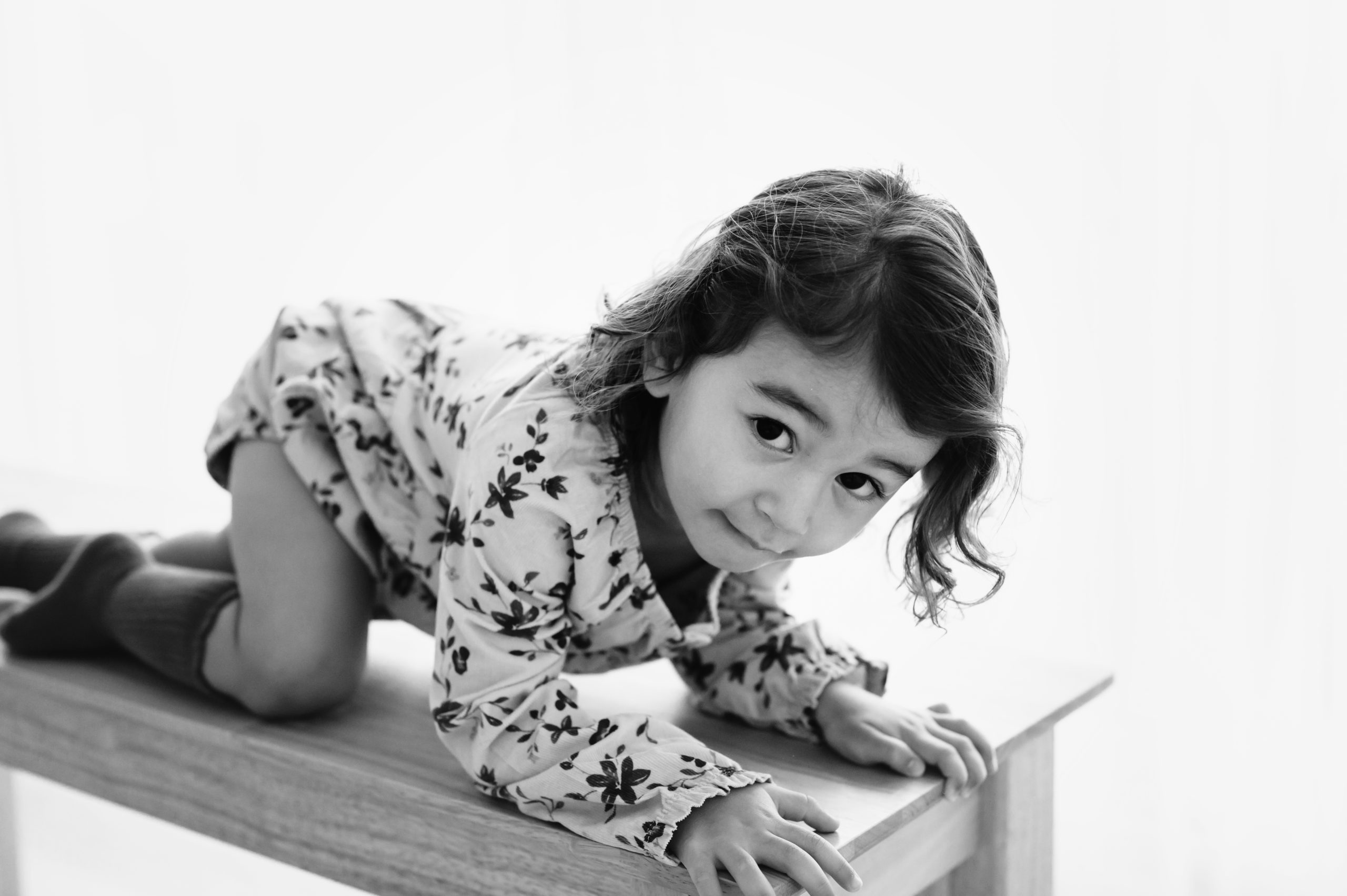 a black and white backlit phot of a young girl crawling across a wooden bench and looking up at the camera during a studio milestone photoshoot
