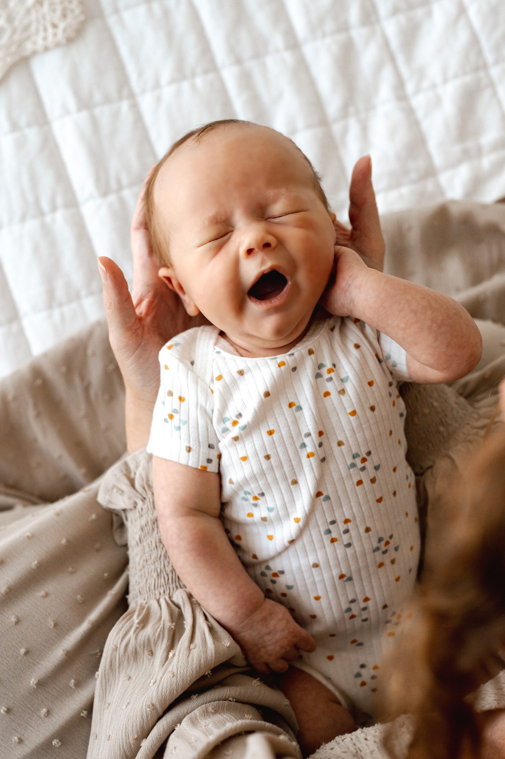 a newborn baby boy yawning and touching his cheek while his mom holds him in her hands during a newborn photoshoot