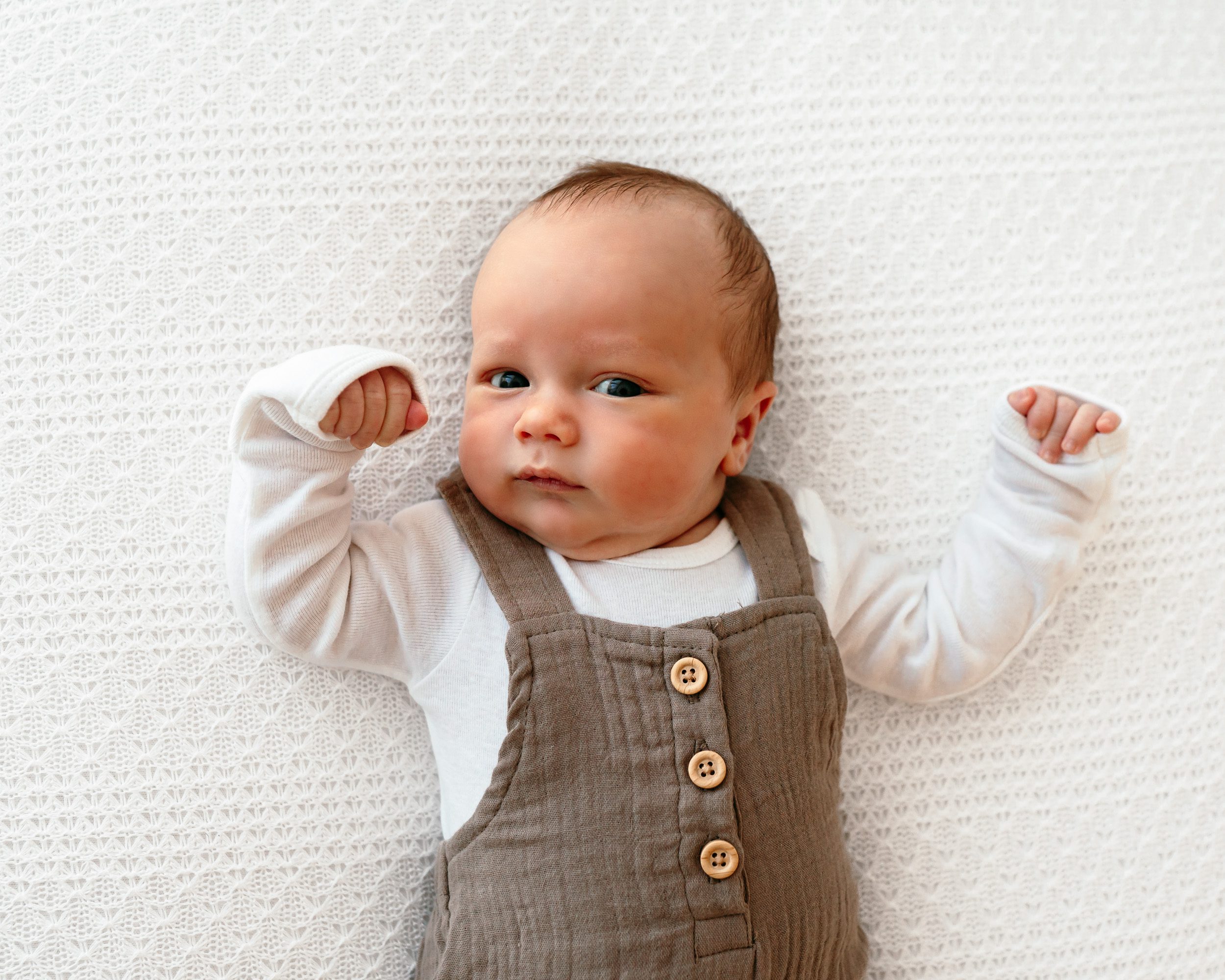 a baby boy in brown overalls laying on a white backdrop and looking up toward the camera during a newborn photoshoot
