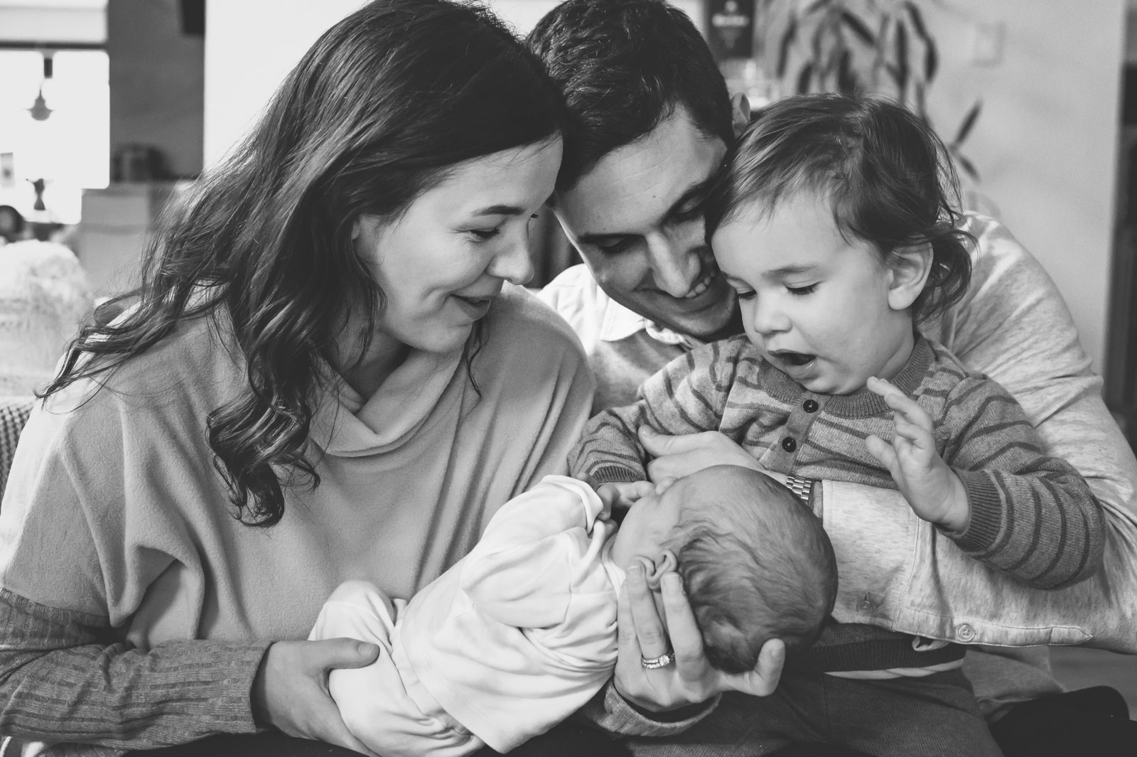 a black and white picture of a mom, dad, and older brother smiling down at their newborn baby boy as mom holds him in her arms during an in home newborn photoshoot