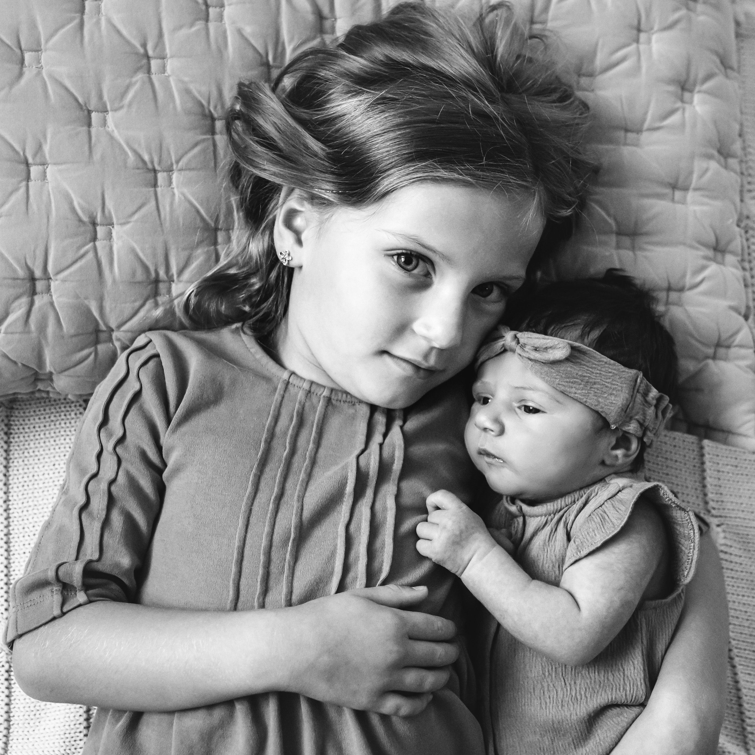 a black and white picture of a girl and her newborn baby sister laying on the floor and snuggling together as big sister looks up at the camera during an in home newborn photo session