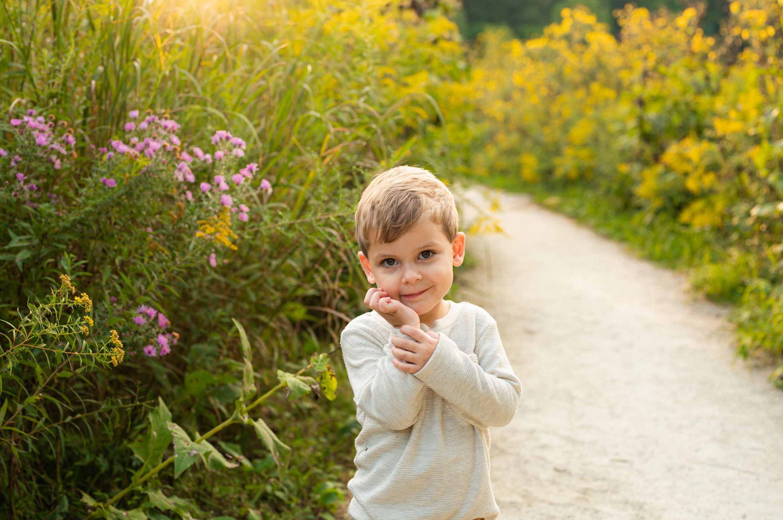 a little boy standing on a path surrounded by yellow wildflowers and holding his hand up to his face during a family photo session