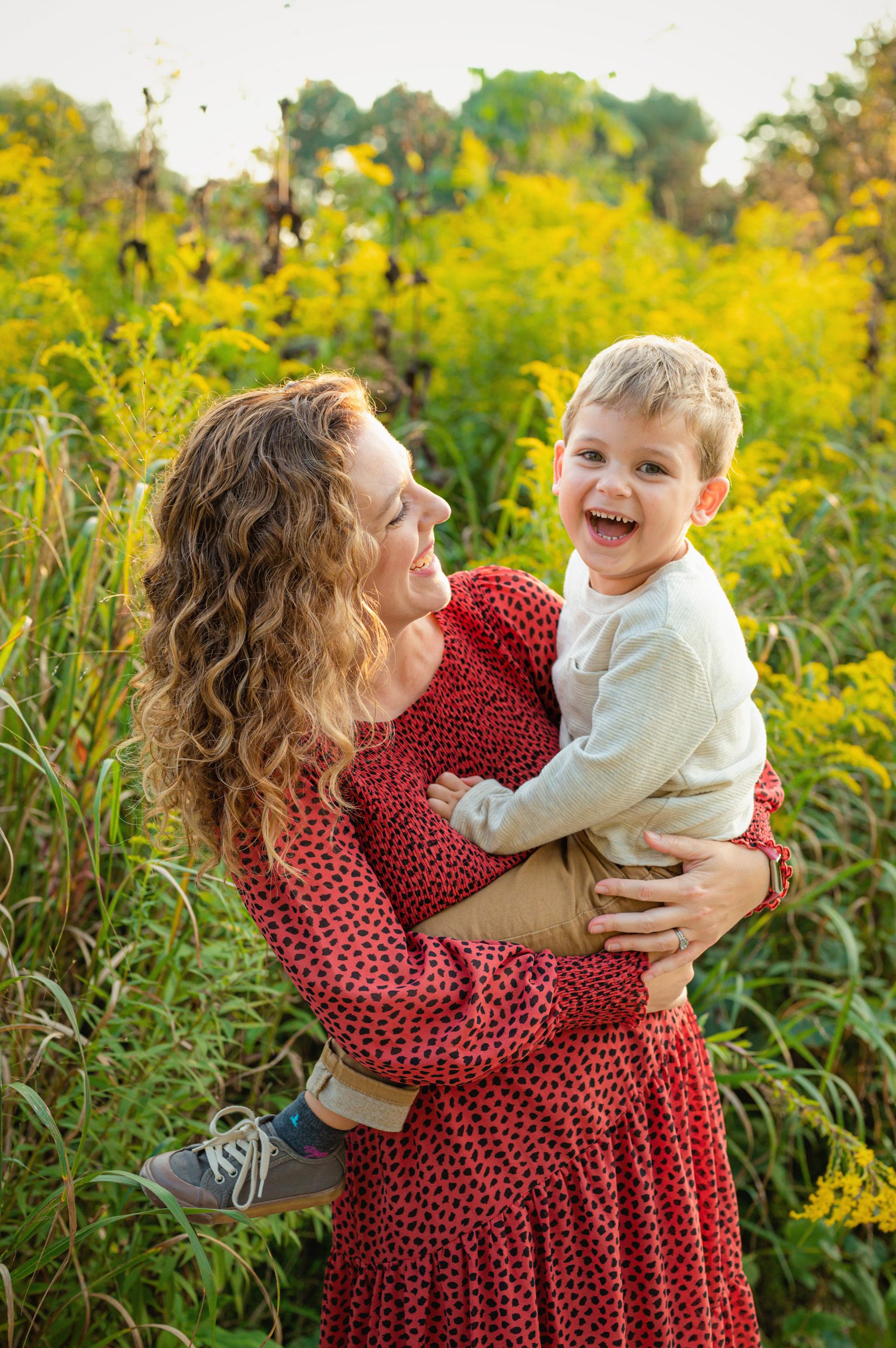 a mom standing in a field of yellow wildflowers and holding her young son in her arms as he smiles at the camera during a West Chester family photo session