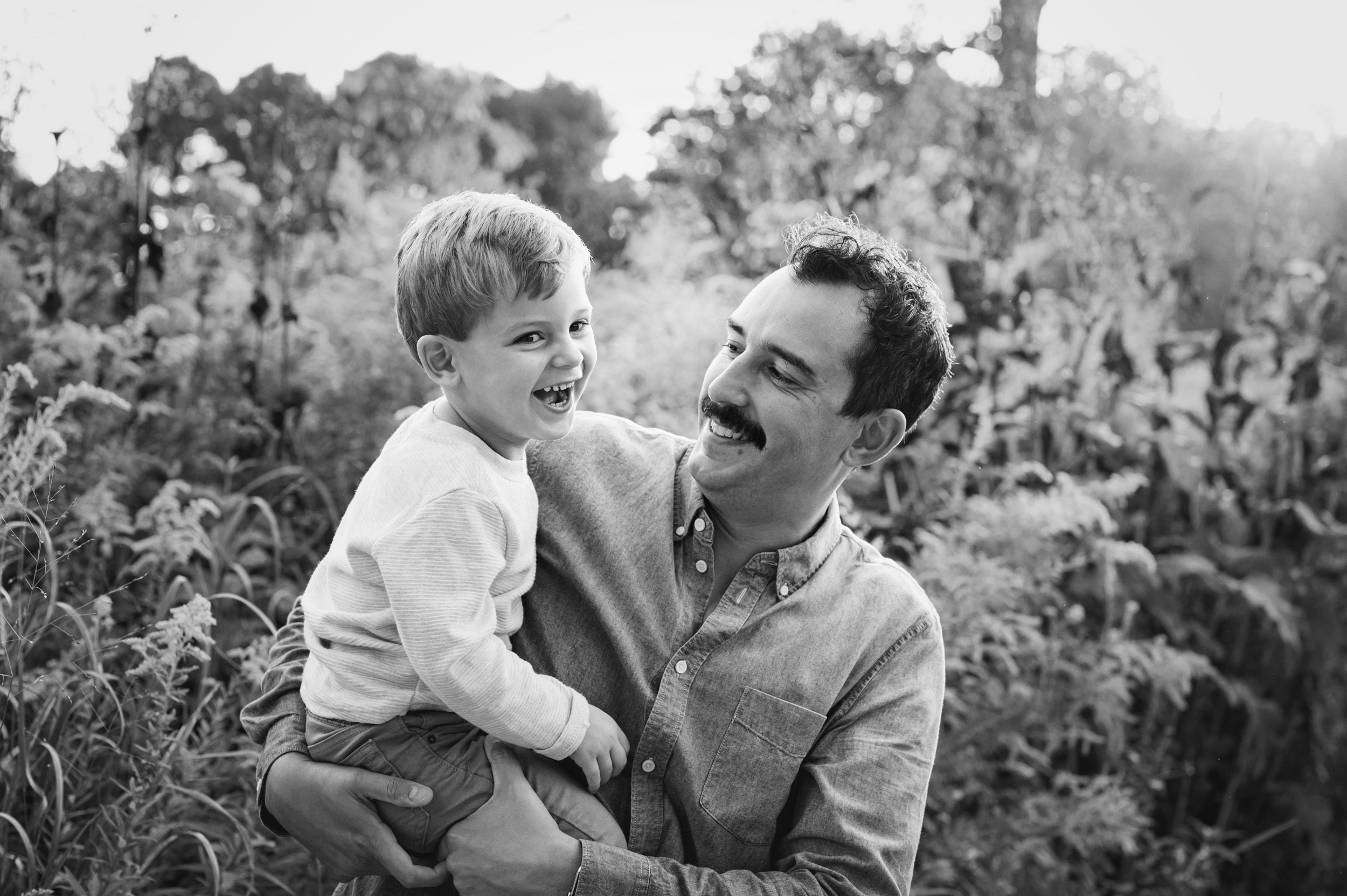 a black and white picture of a dad holding his young son in his arms as they both laugh during a family photoshoot