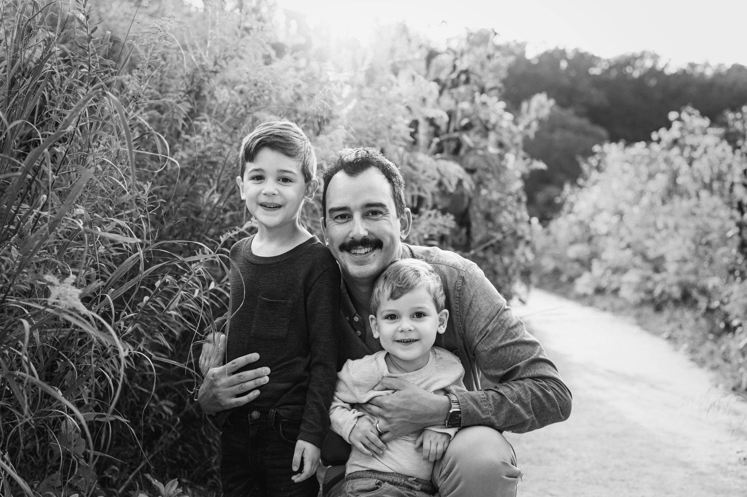 a black and white picture of a dad kneeling in a field of flowers with his arms around his two young sons during a family photo session