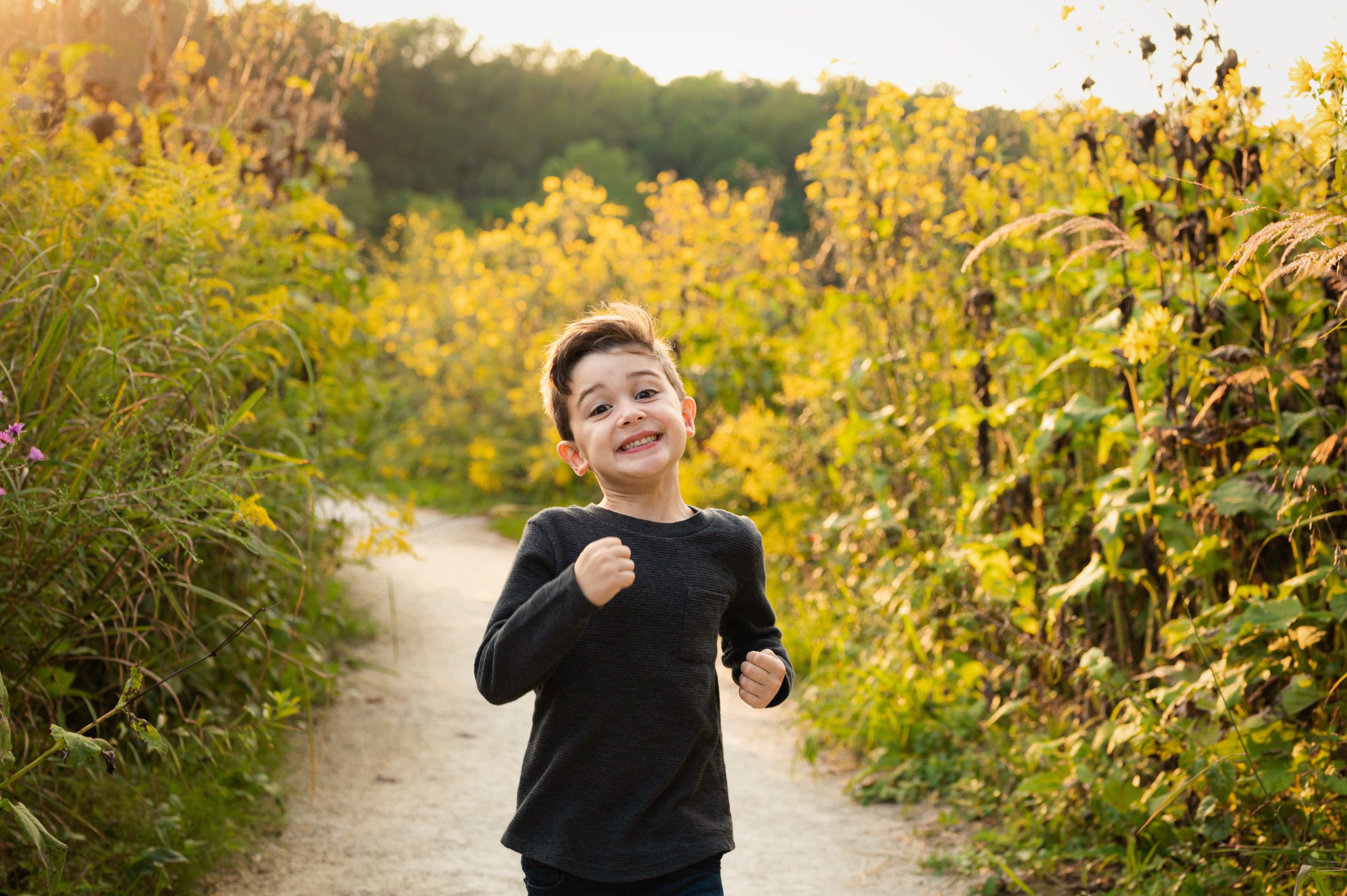 a young boy running down a path surrounded by yellow wildflowers with a huge smile on his face during a family photoshoot