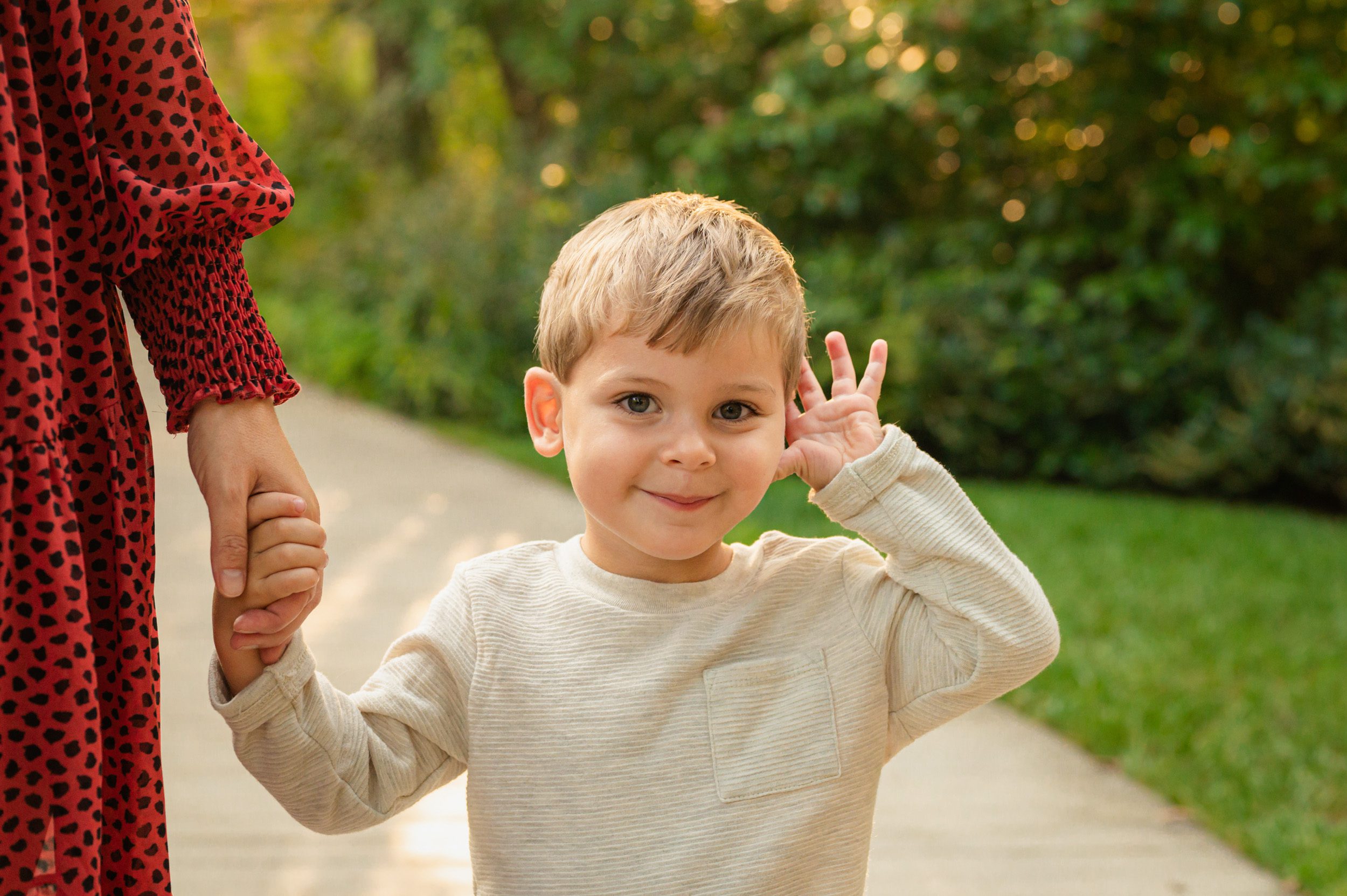 a young boy holding his mom's hand and smiling shyly at the camera as he holds his hand up to his face during a Longwood Gardens family photo session