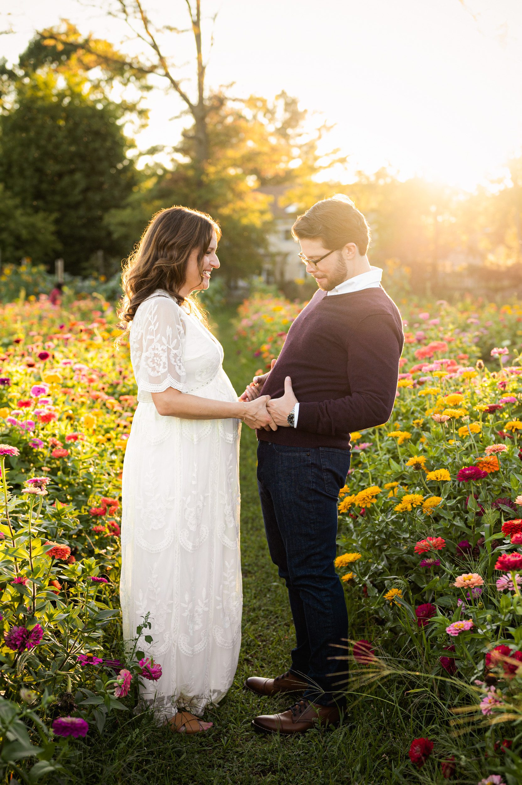 an expecting couple standing in a field of colorful wildflowers and laughing as the dad sticks out his belly during a maternity photoshoot