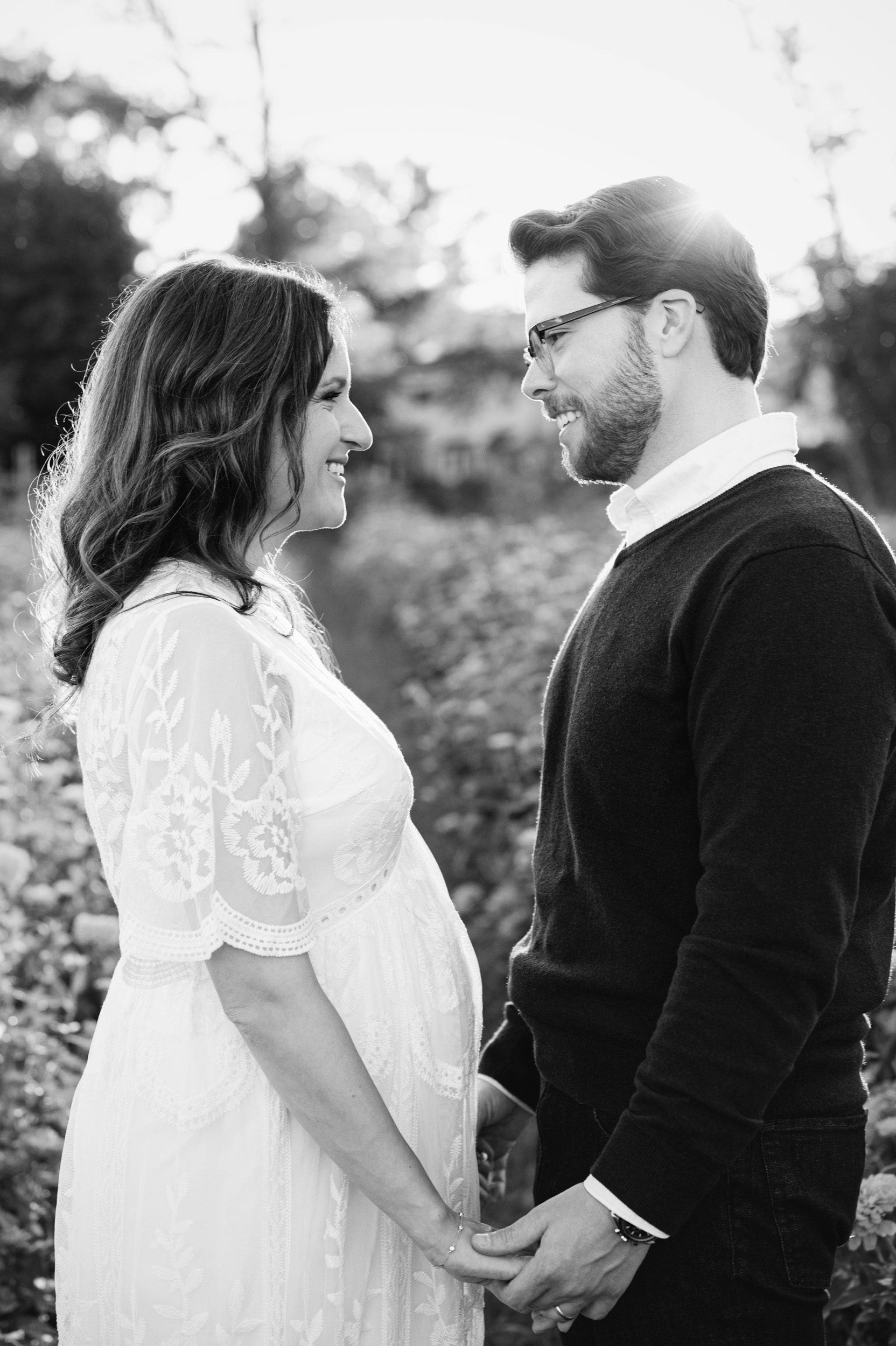 a black and white picture of an expecting couple holding hands and smiling at each other while the sunset shines in the background during a maternity photoshoot