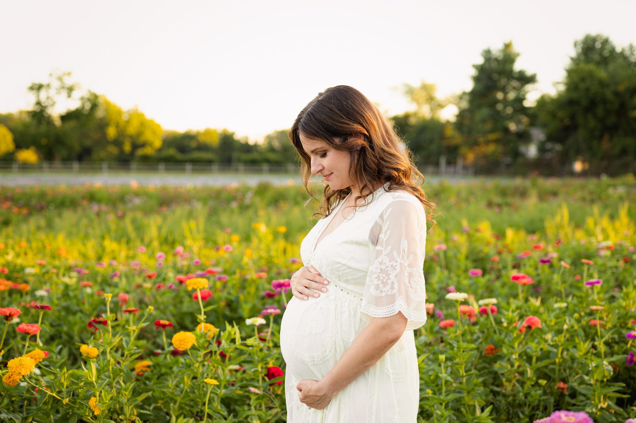 an expecting mother in a white dress standing in a field of colorful wildflowers and smiling down at her belly during a Philadelphia maternity photoshoot