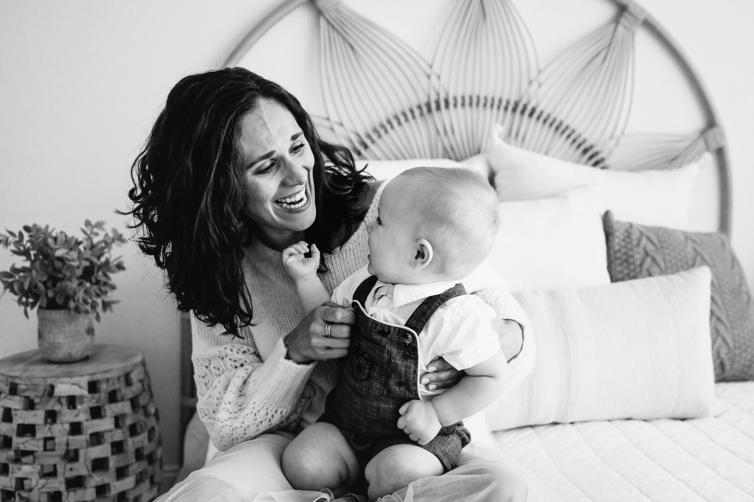 a black and white picture of a mom sitting on a bed with her baby boy on her lap as he reaches back toward her and they both laugh during a baby milestone photo session