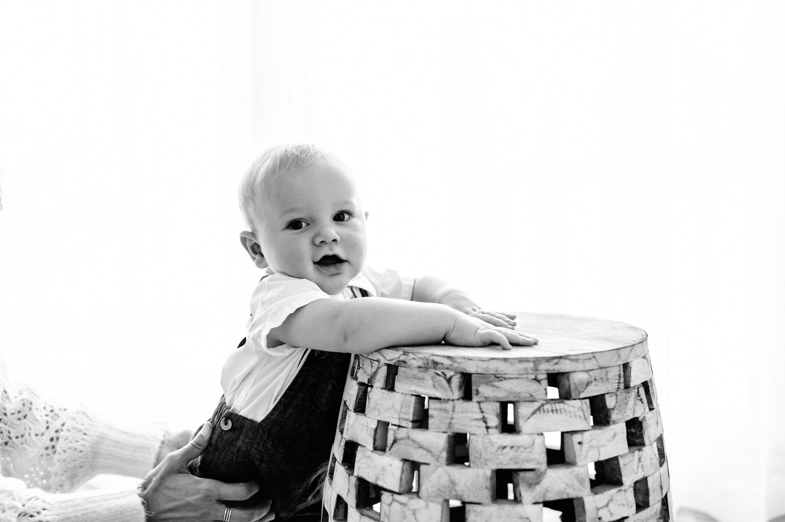 a black and white backlit picture of a baby boy holding onto a barrel and looking at the camera while his mom holds him steady with her hands during a baby milestone photoshoot