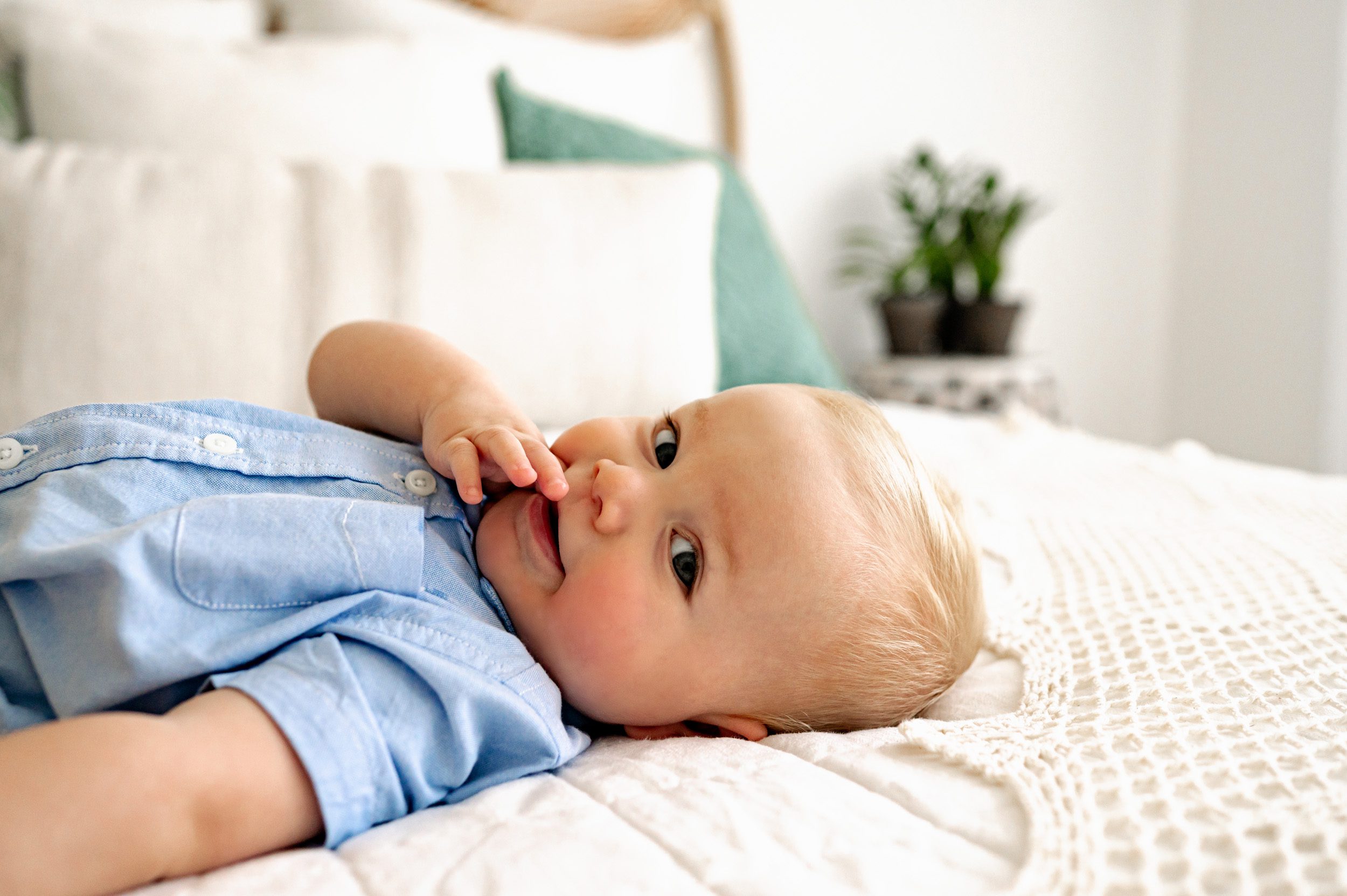 a baby boy laying on a bed and looking shyly toward the camera while he sucks on his finger during a baby milestone photoshoot