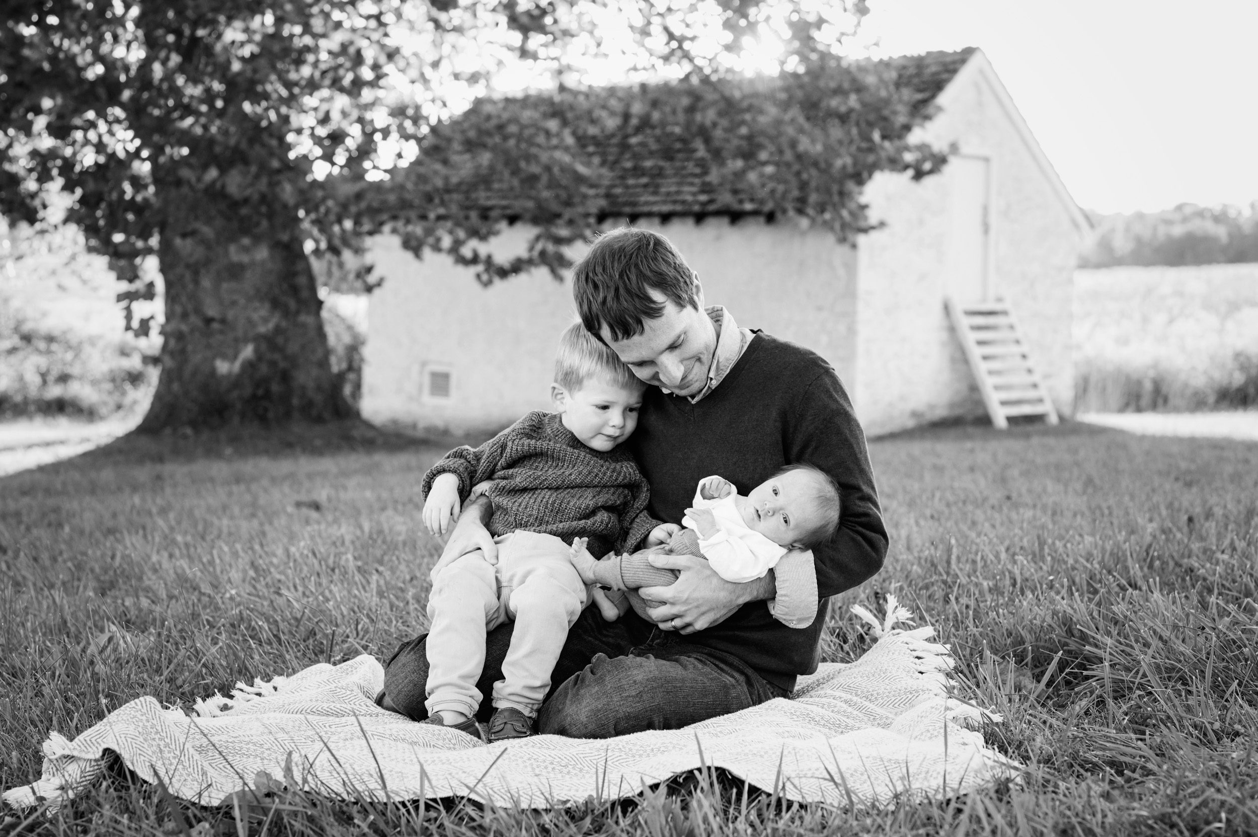 a black and white picture of a dad sitting in the grass with his two sons on his lap and smiling down at his baby boy during a King of Prussia family photoshoot
