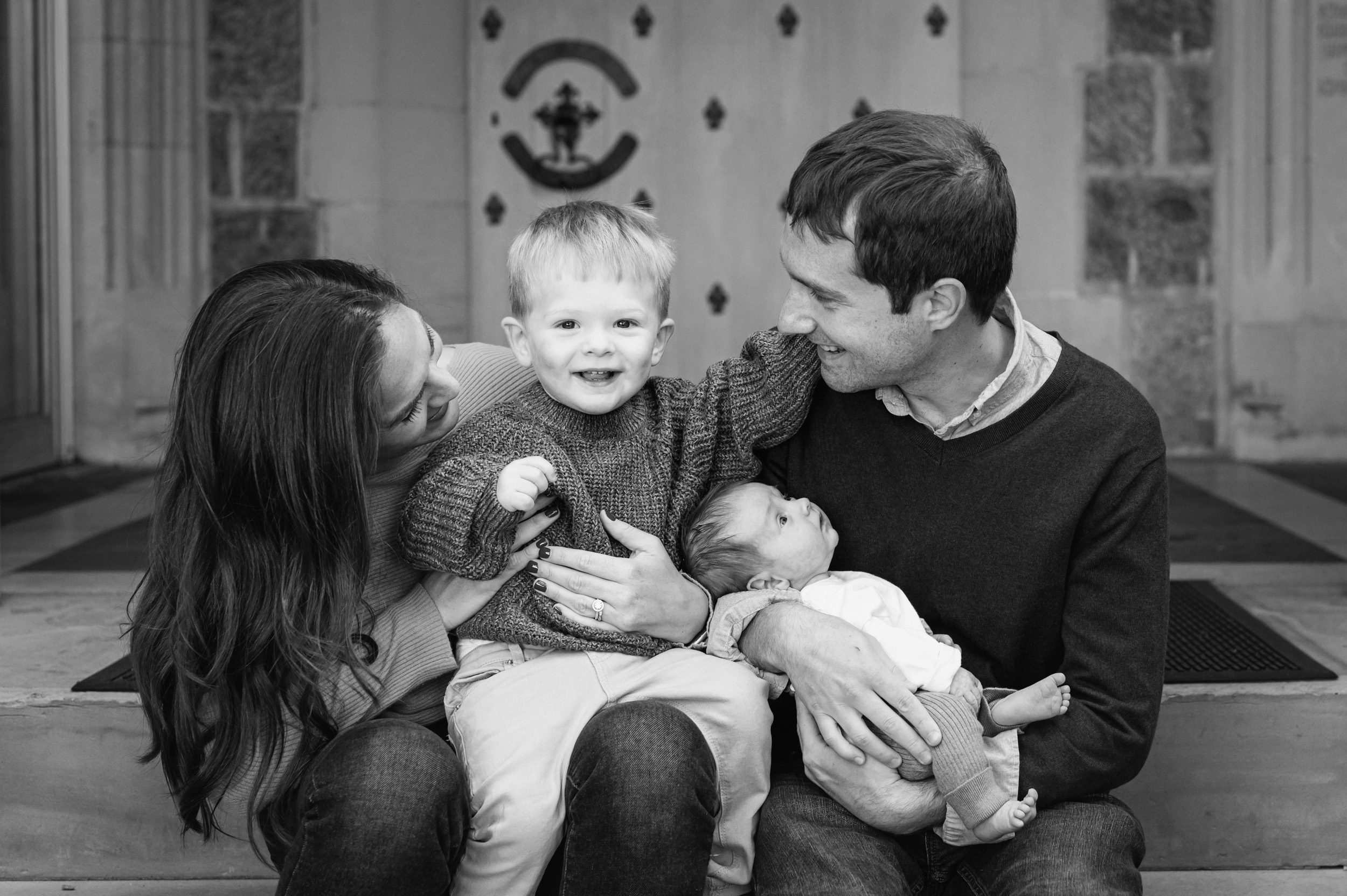 a black and white picture of a young boy sitting on his parents lap and touching his dad's cheeks while his baby brother looks up at him during a King of Prussia family photoshoot