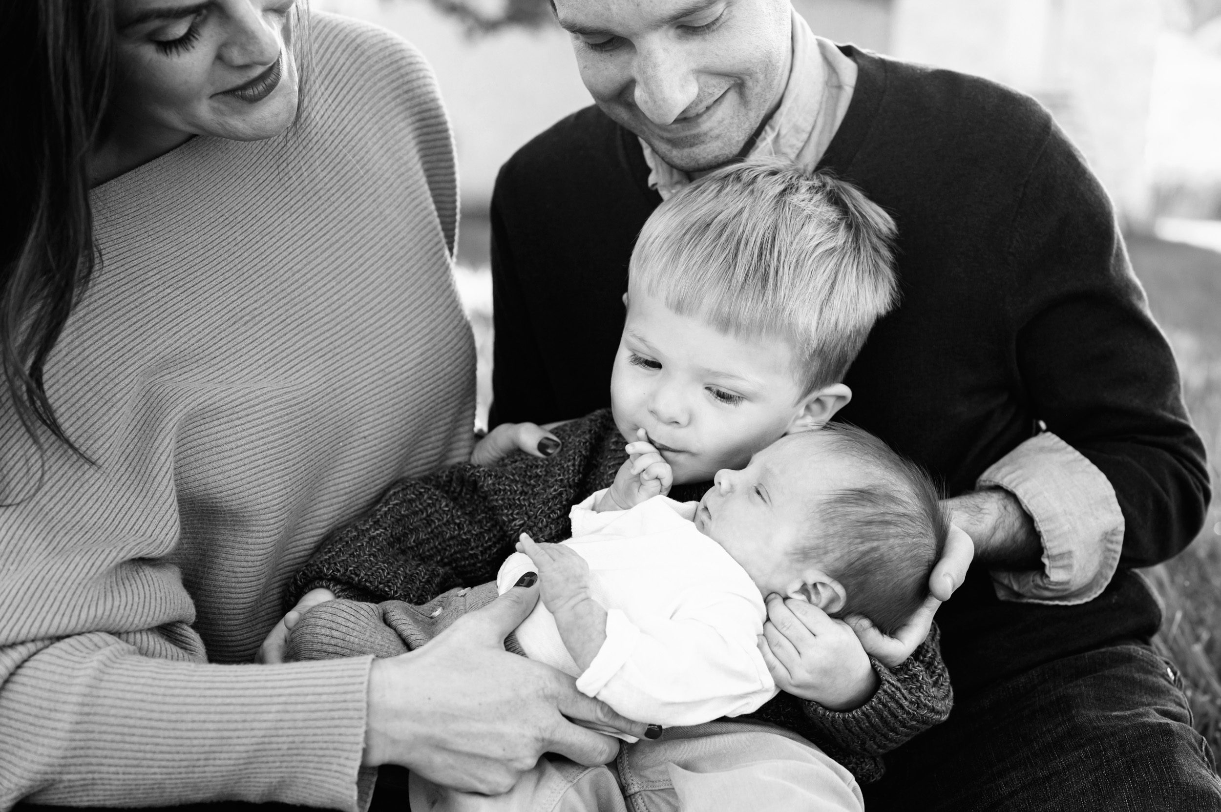 a black and white picture of a new big brother sitting on his parents' lap and kissing his new baby brother's hand during a King of Prussia family photoshoot