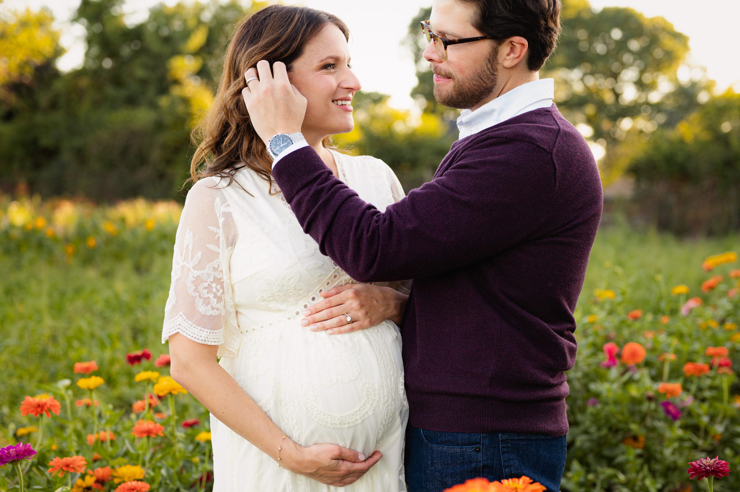 an expecting mom standing in a flower field cradling her belly as her husband gently brushes the hair out of her face during a maternity photoshoot