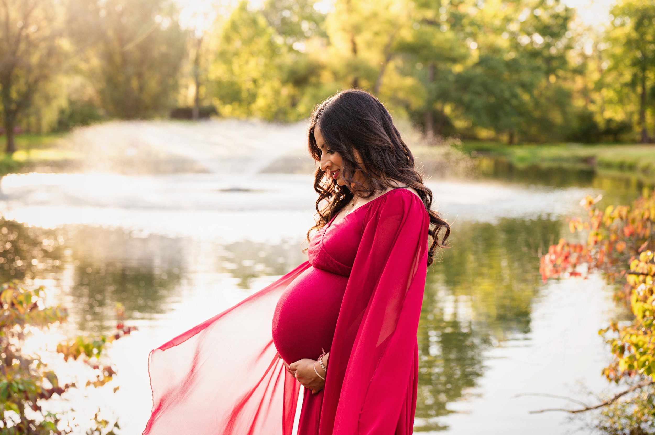 an expecting mother in a red dress standing in front of a pond smiling down at her belly as the sun shines through a fountain in the background during a maternity photo session