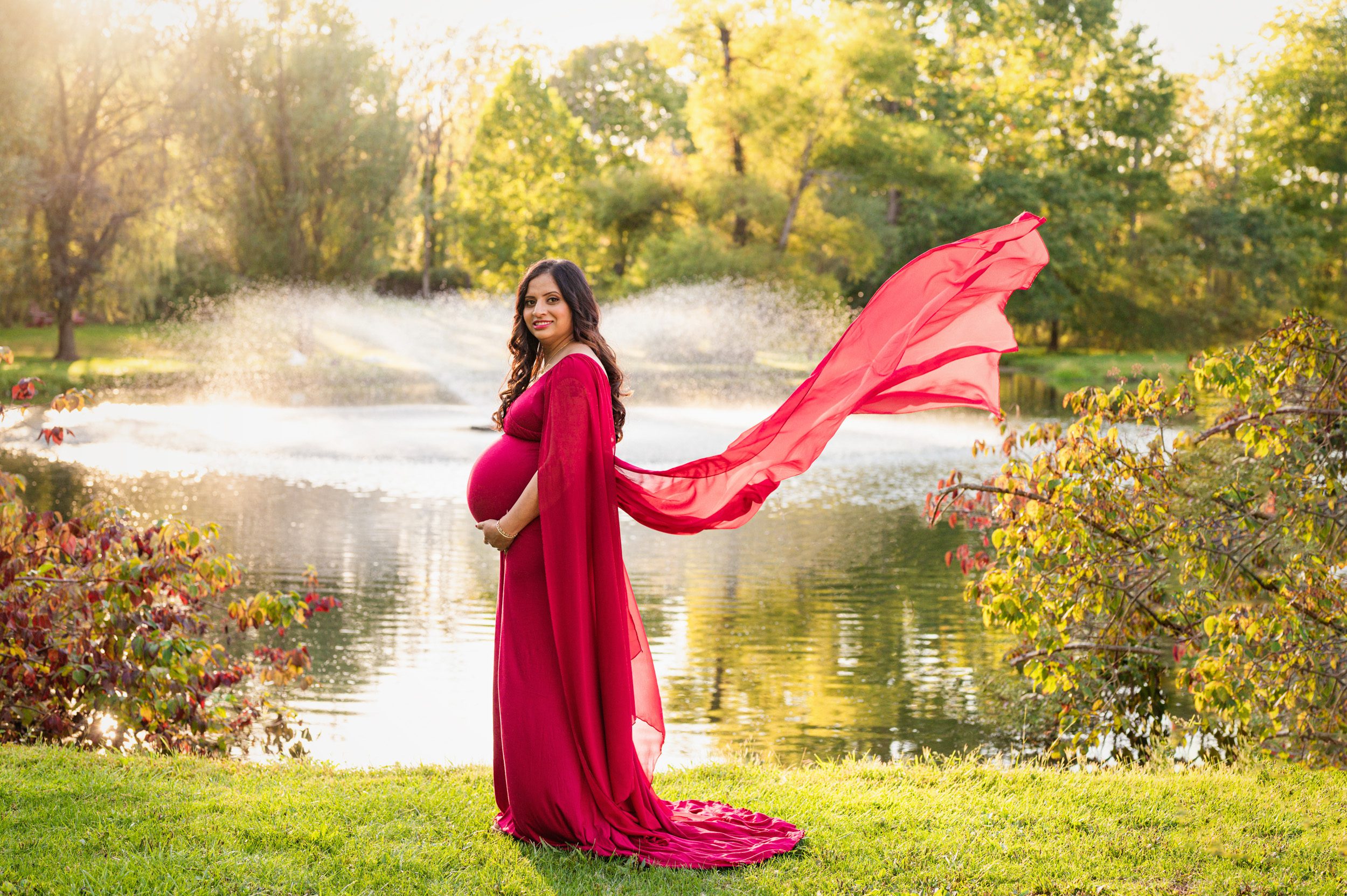 an expecting mother in a red dress standing in front of a pond with the train of her dress flowing out behind her during a maternity photoshoot