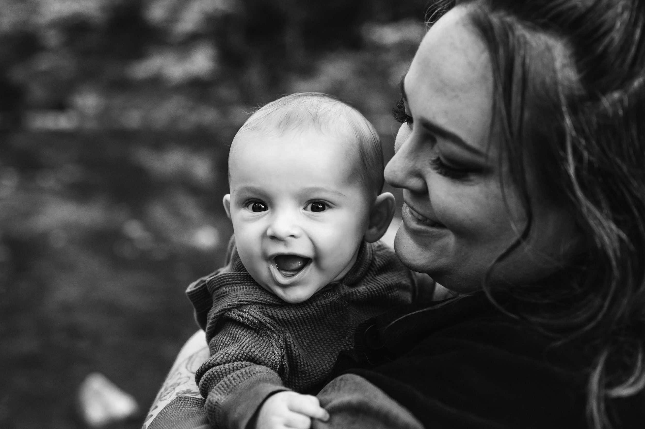 a black and white picture of a baby boy looking over his mom's shoulder and laughing as she smiles at him adoringly during a family photo session
