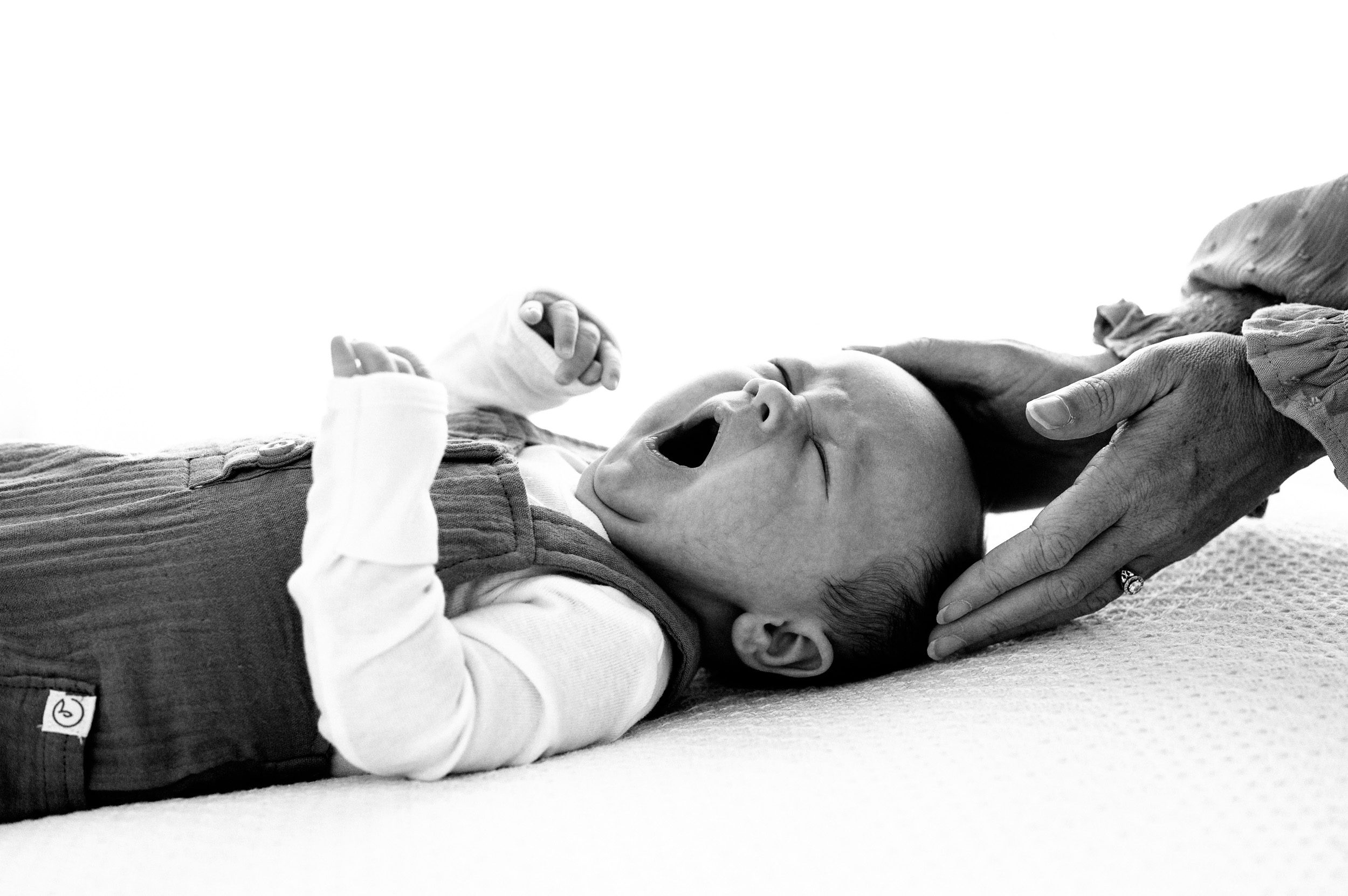 a black and white backlit picture of a newborn baby boy laying on a white blanket and yawning while his mom cradles his head in her hands during a newborn photoshoot