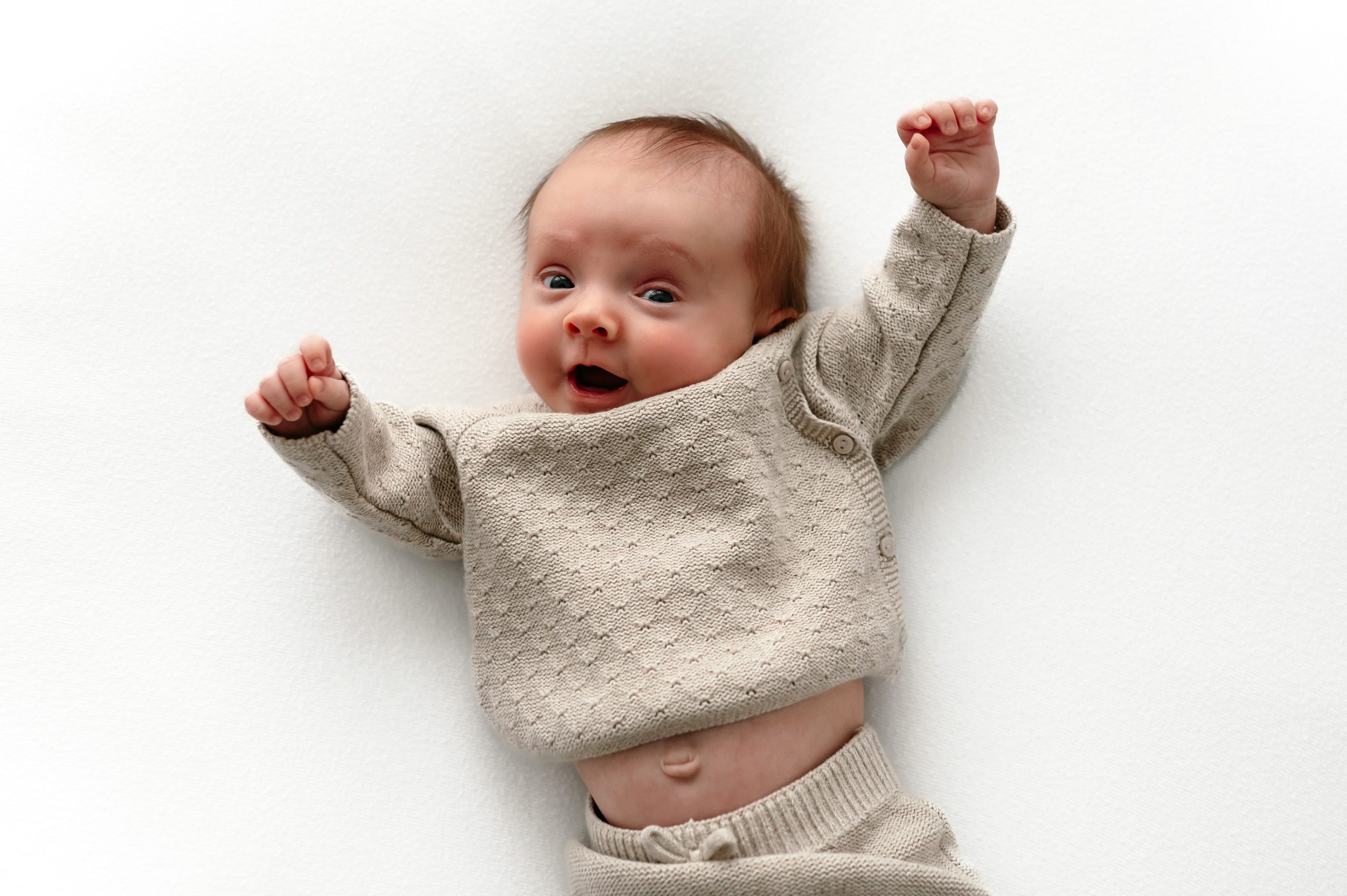 a newborn baby boy laying on a white backdrop and lifting his arms above his head as he looks at the camera during a newborn photoshoot