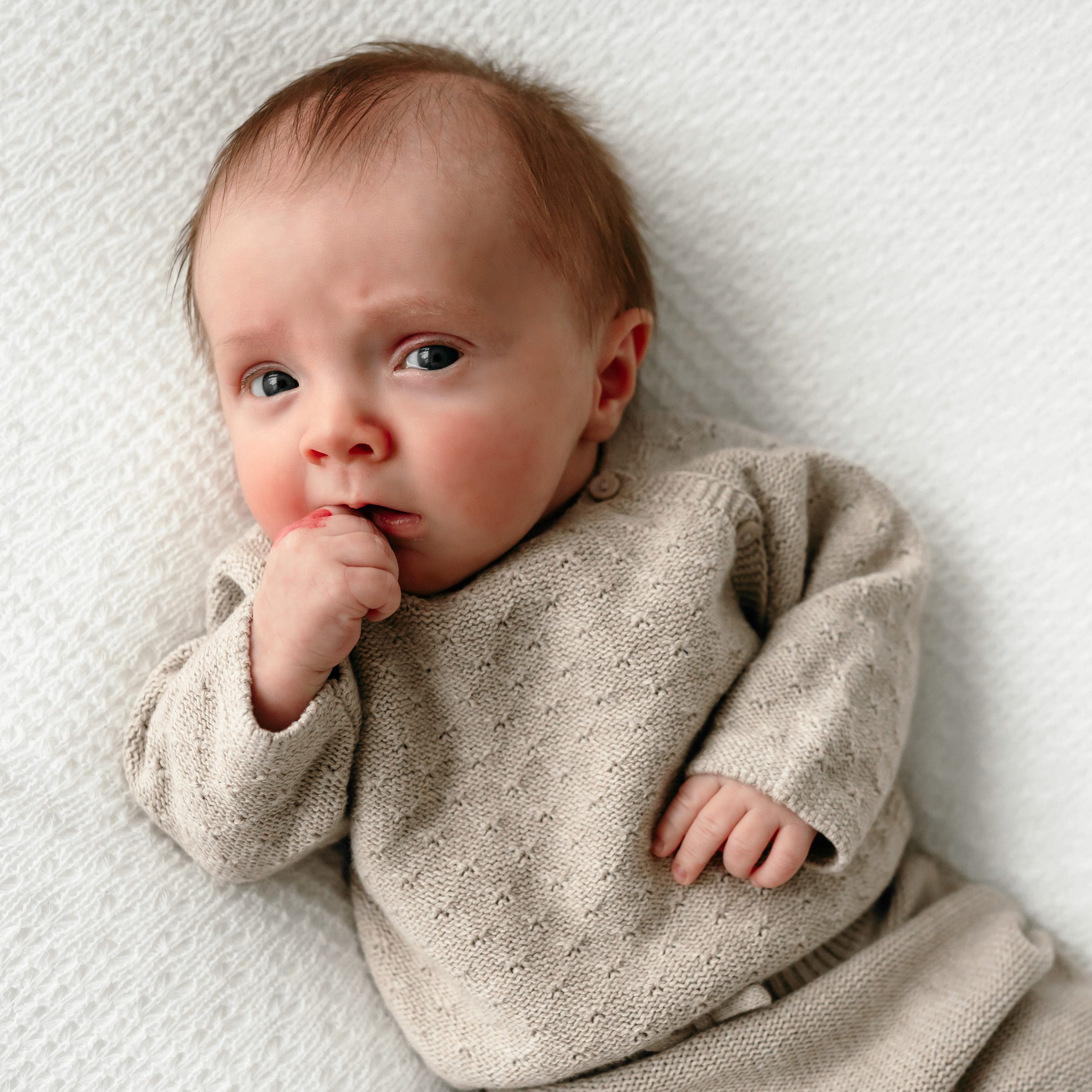a newborn boy laying on a white backdrop holding his hand to his lips and looking up toward the camera with a concerned expression on his face during a newborn photoshoot 