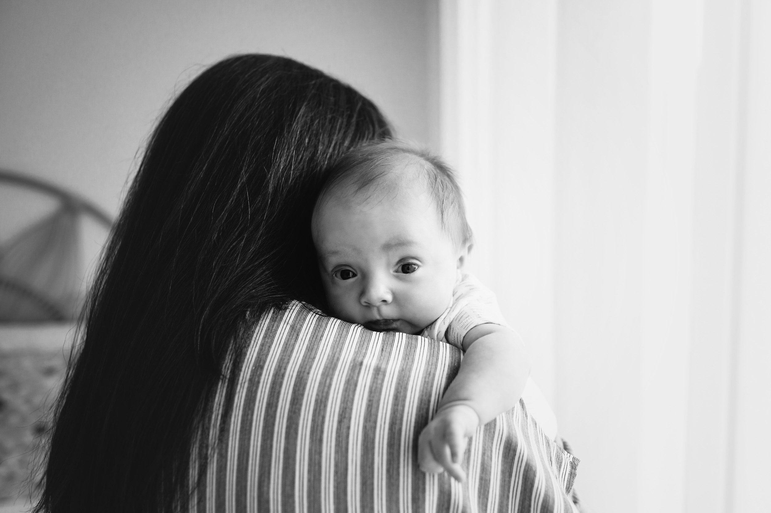 a black and white picture of a baby boy resting his head on his mom's shoulder and looking at the camera during a newborn photoshoot