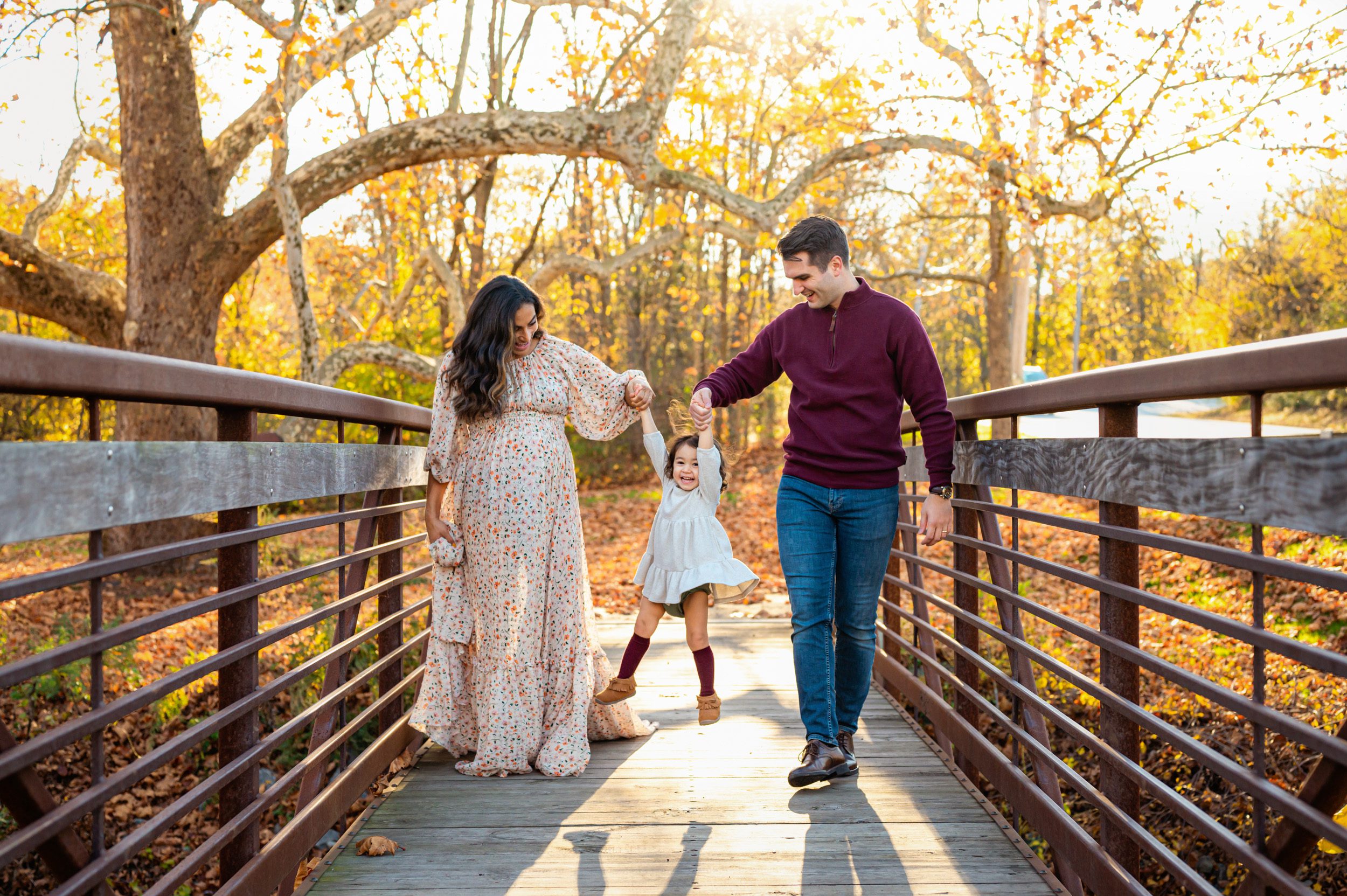 a little girl holding her mom and dad's hands as they swing her up in the air and laugh while walking across a bridge during a maternity photoshoot