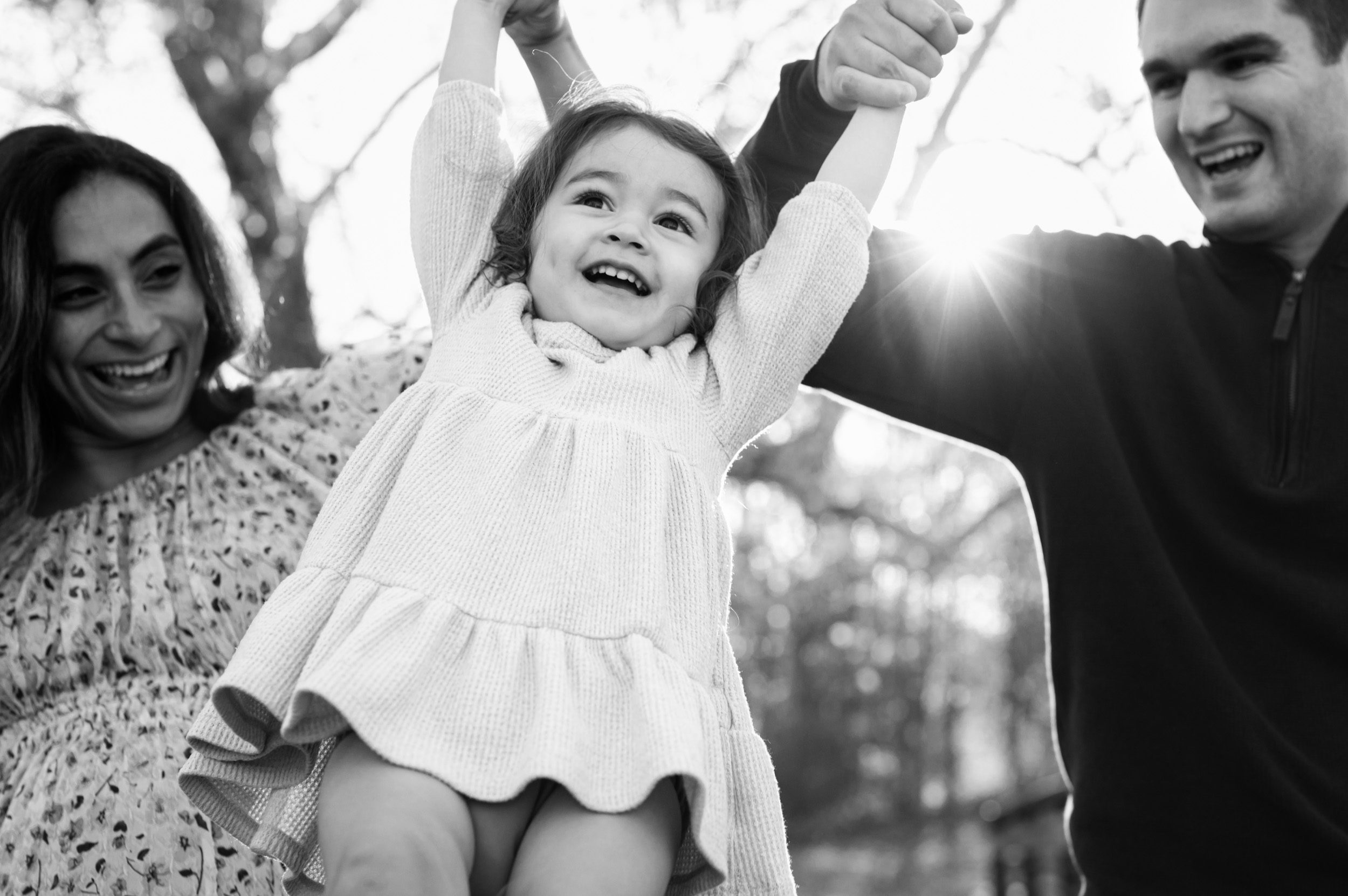 a close up black and white picture of a little girl holding her mom and dad's hands and swinging high up in the air with the sunset shining through in the background during a maternity photo session