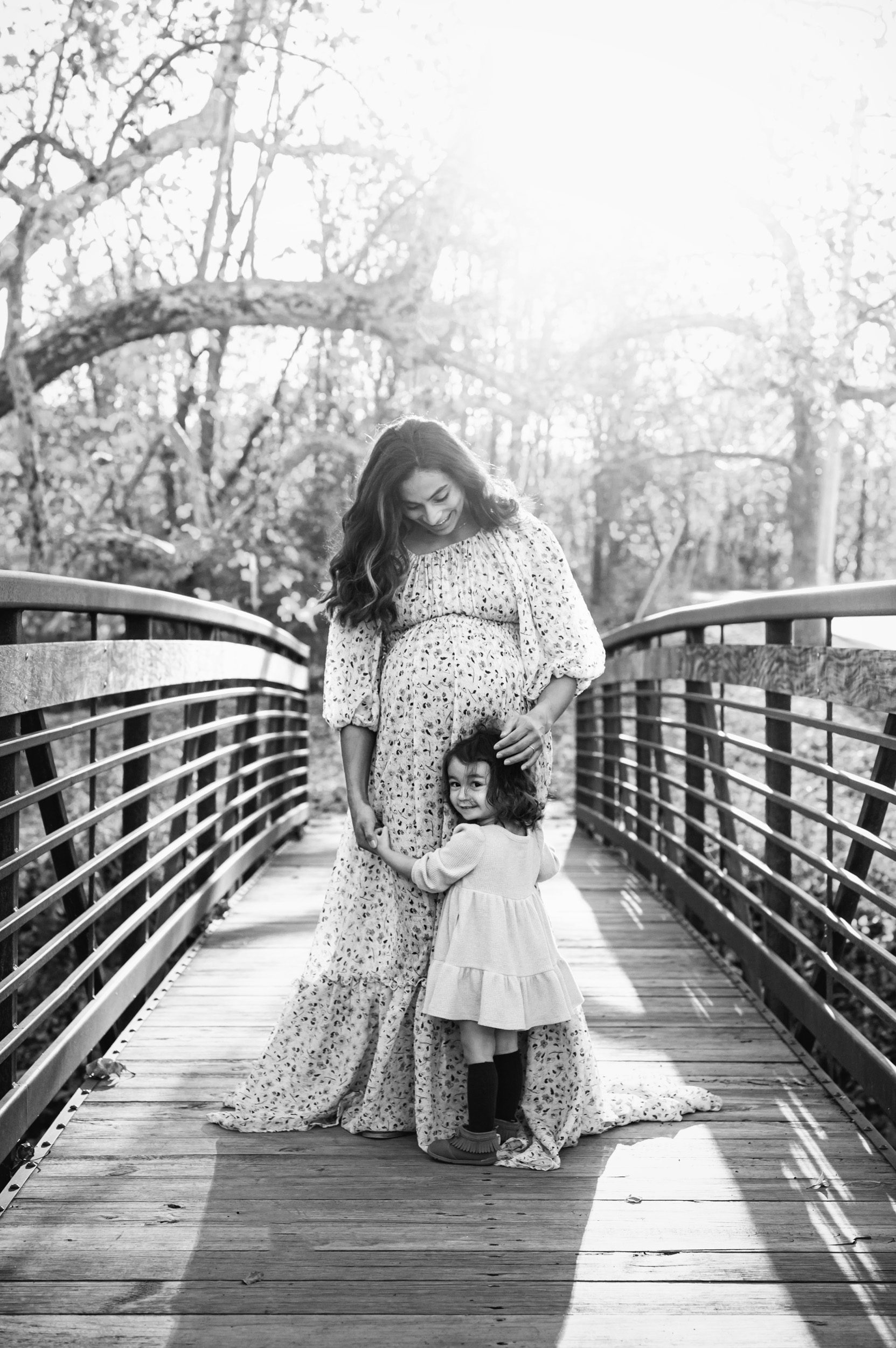 a black and white picture of an expecting mom standing on a bridge hugging her daughter during a maternity photoshoot