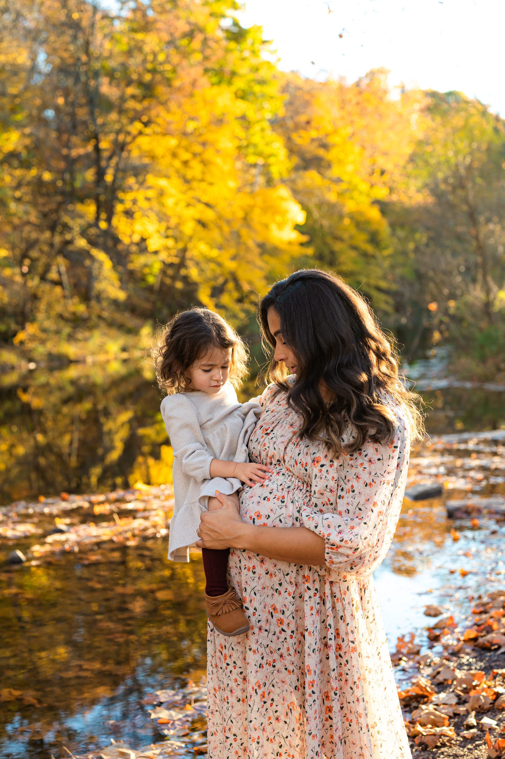 an expecting mother standing in front of a creek holding her daughter as they both look down and touch her belly during a maternity photoshoot