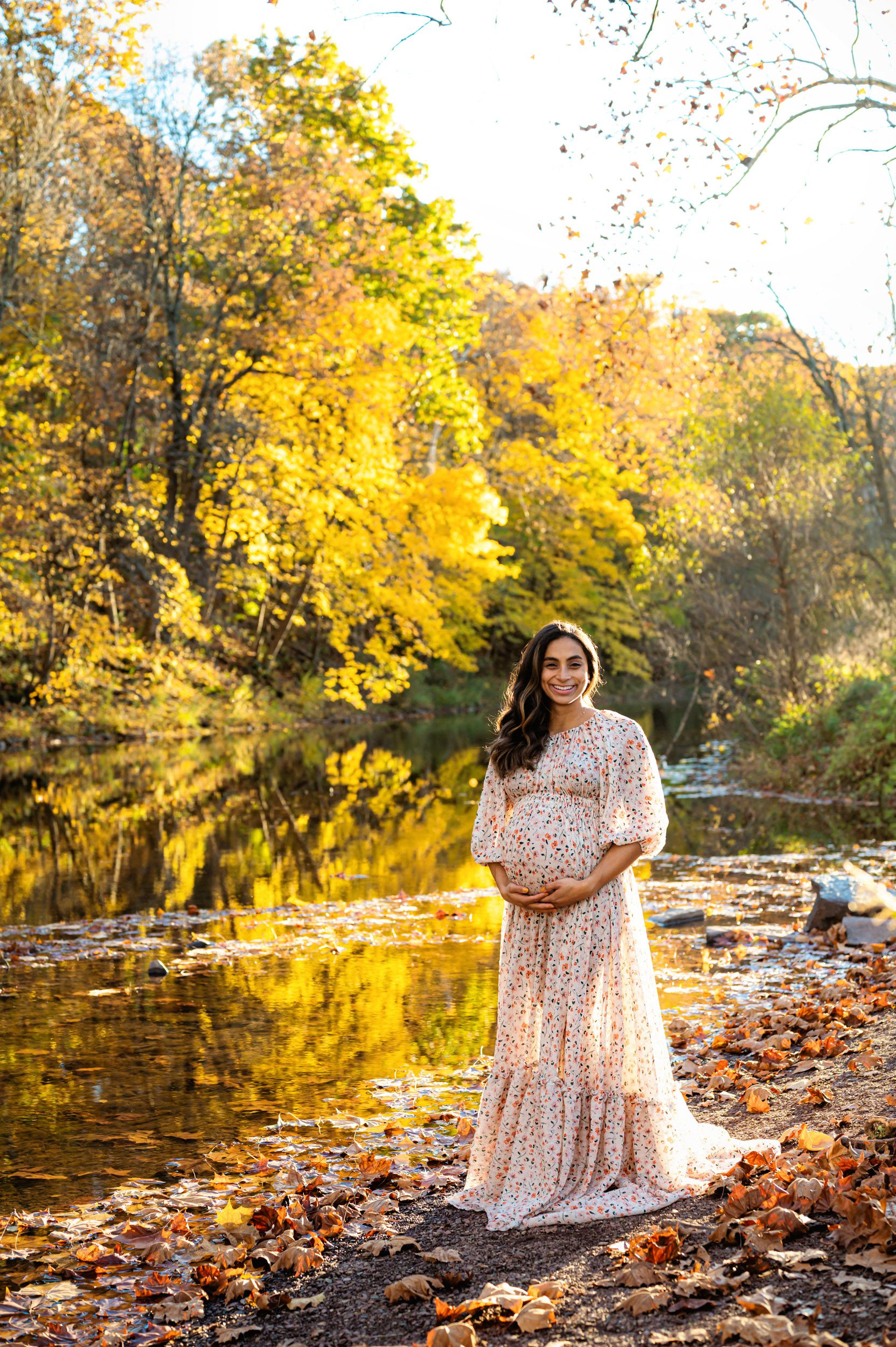 an expecting mother standing in front of a creek cradling her belly with colorful autumn leaves reflecting off the water in the background during a maternity photoshoot