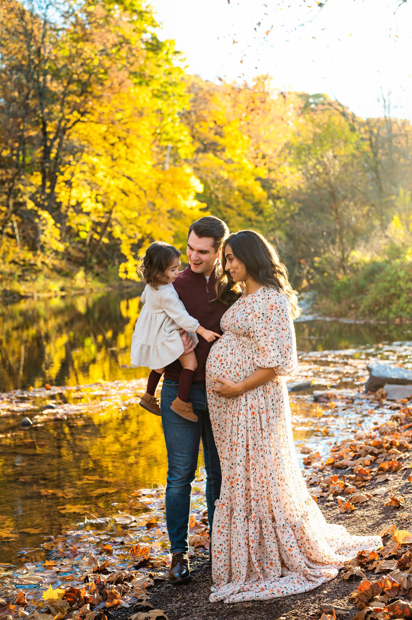 a pregnant mom standing on the edge of a creek while her young daughter points at her belly and the colorful fall leaves reflect off the water in the background during a maternity photoshoot