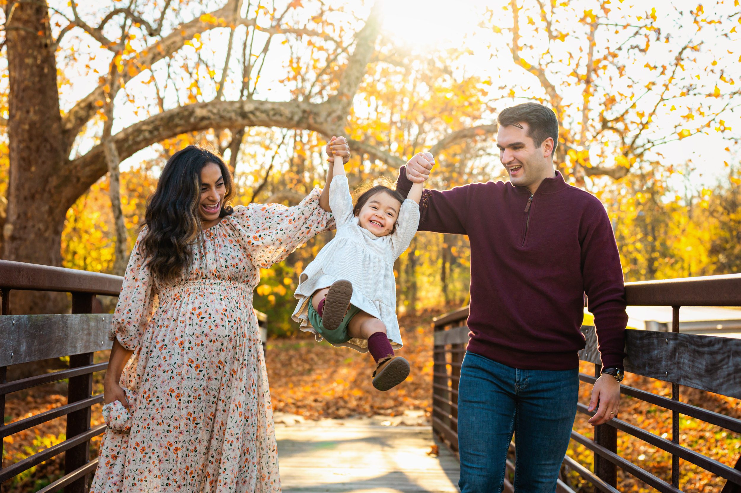 a little girl holding her mom and dad's hands as they swing her high up in the air and laugh while walking across a bridge during a maternity photoshoot