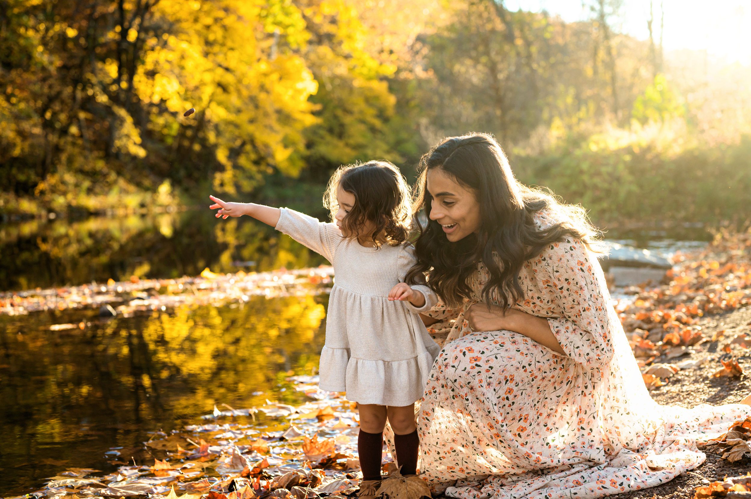 a young girl throwing a stone into a creek with the colorful autumn leaves reflecting off the water in the background during a maternity photo session