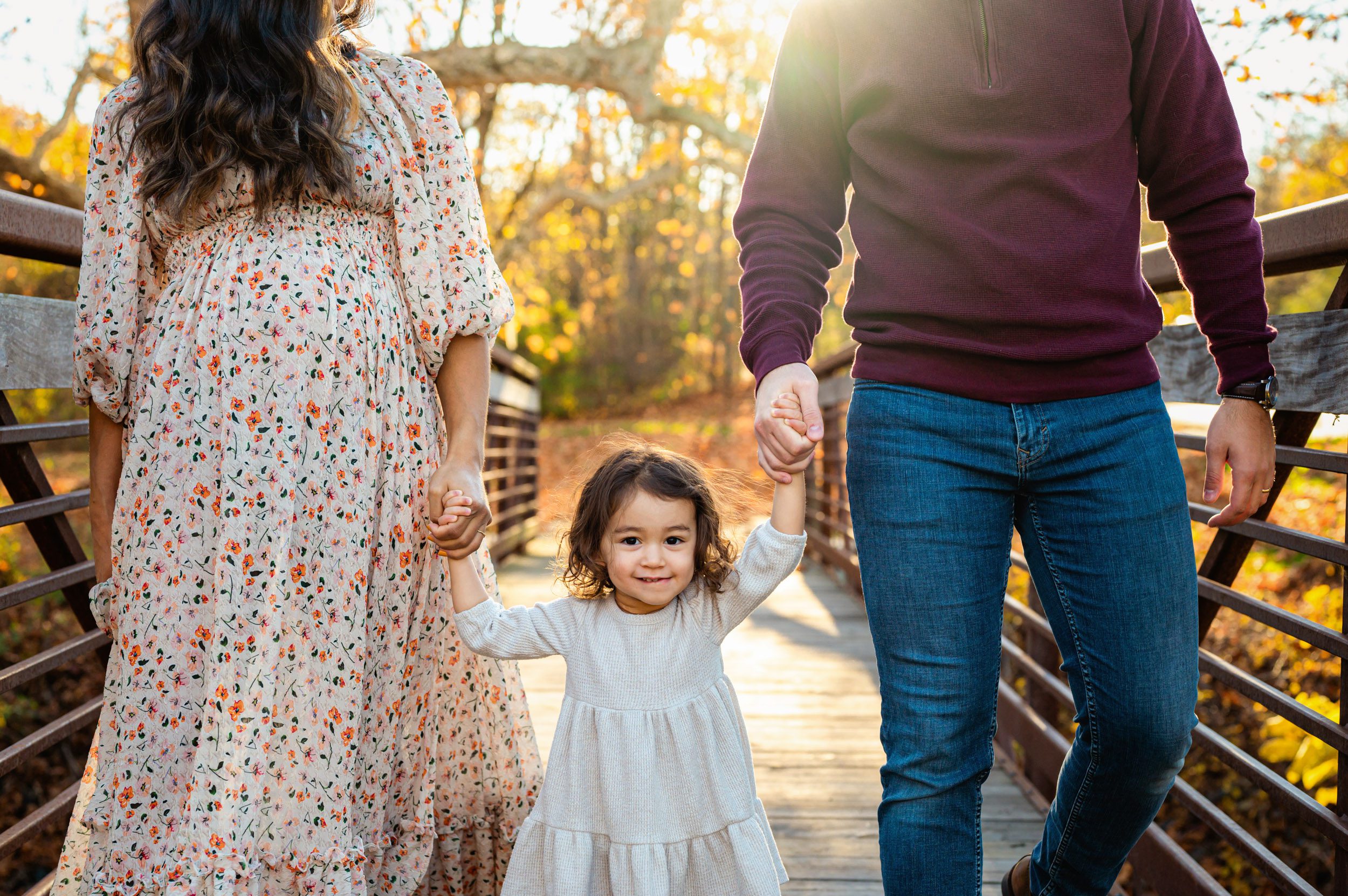 a young girl holding her mom and dad's hands and walking across a bridge as she smiles at the camera during a maternity photo session