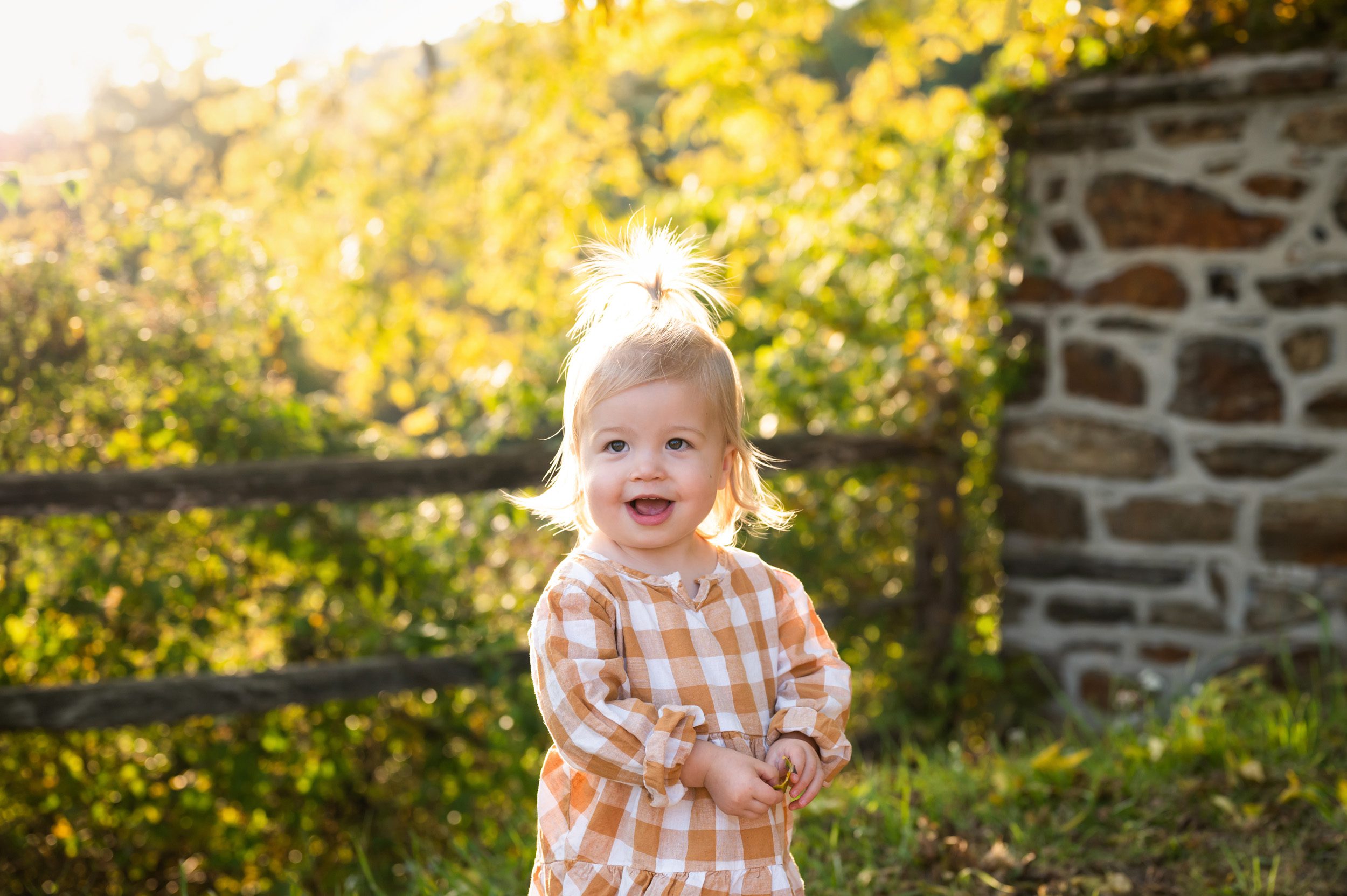 a young girl standing in front of a fence and smiling with the sunlight shining through the colorful fall leaves and lighting up her ponytail during a maternity photoshoot