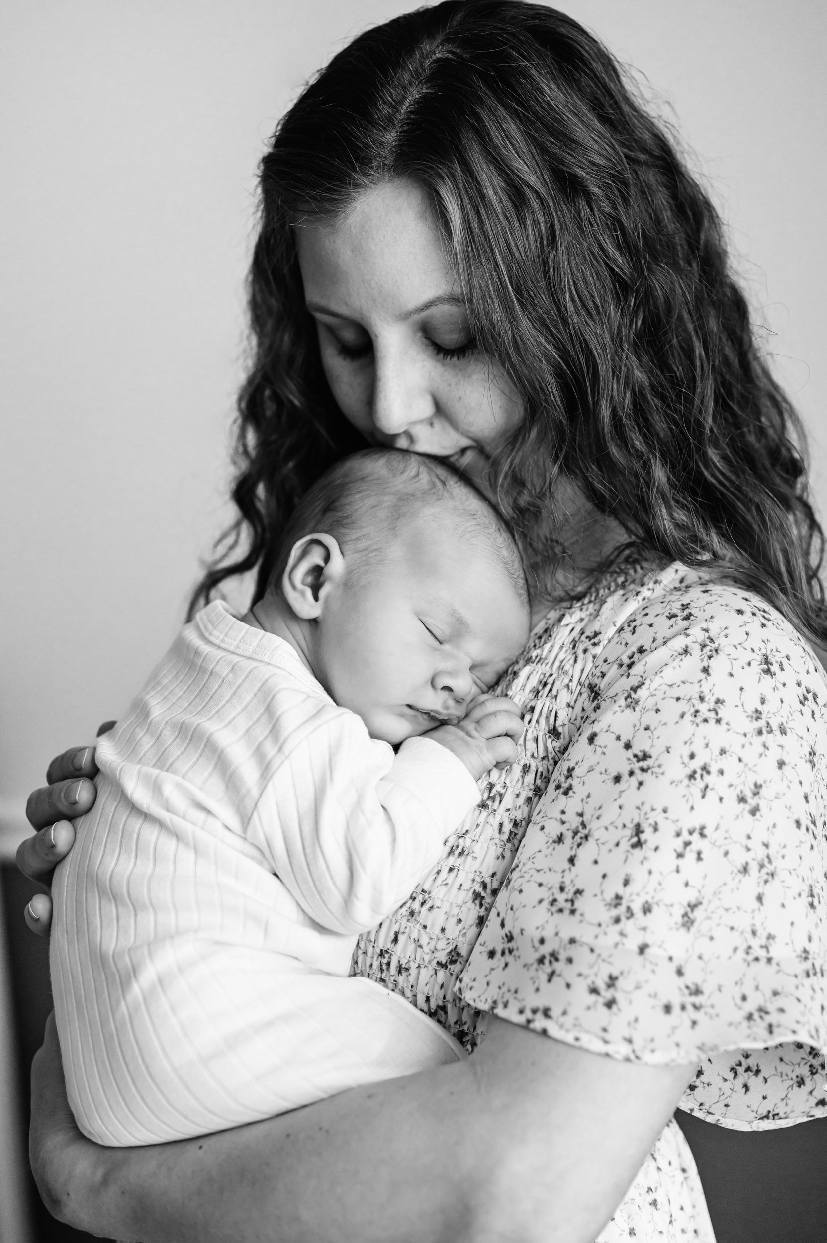 a black and white picture of a mom holding her newborn baby boy against her chest and kissing him on the head during a newborn photography session