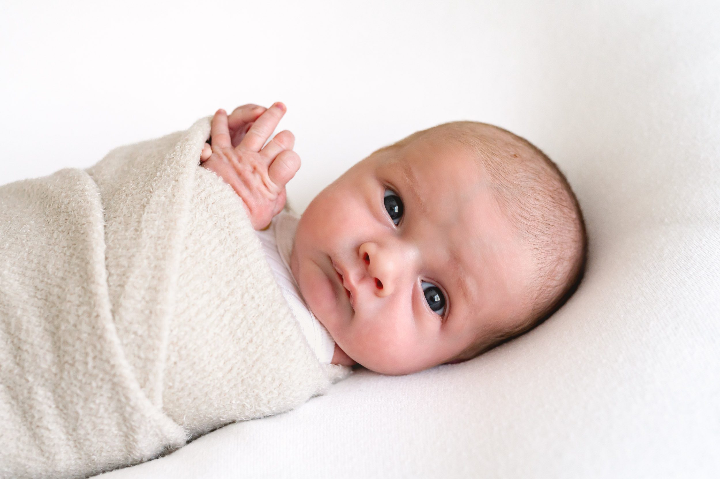newborn baby boy in a white romper and swaddle looking directly at the camera during a newborn baby photography session