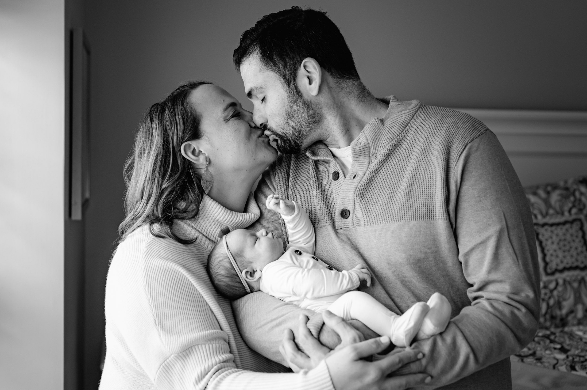 a black and white photo of parents holding their baby girl in their arms and kissing each as their baby girl reaches her hand up toward them during a newborn photo session