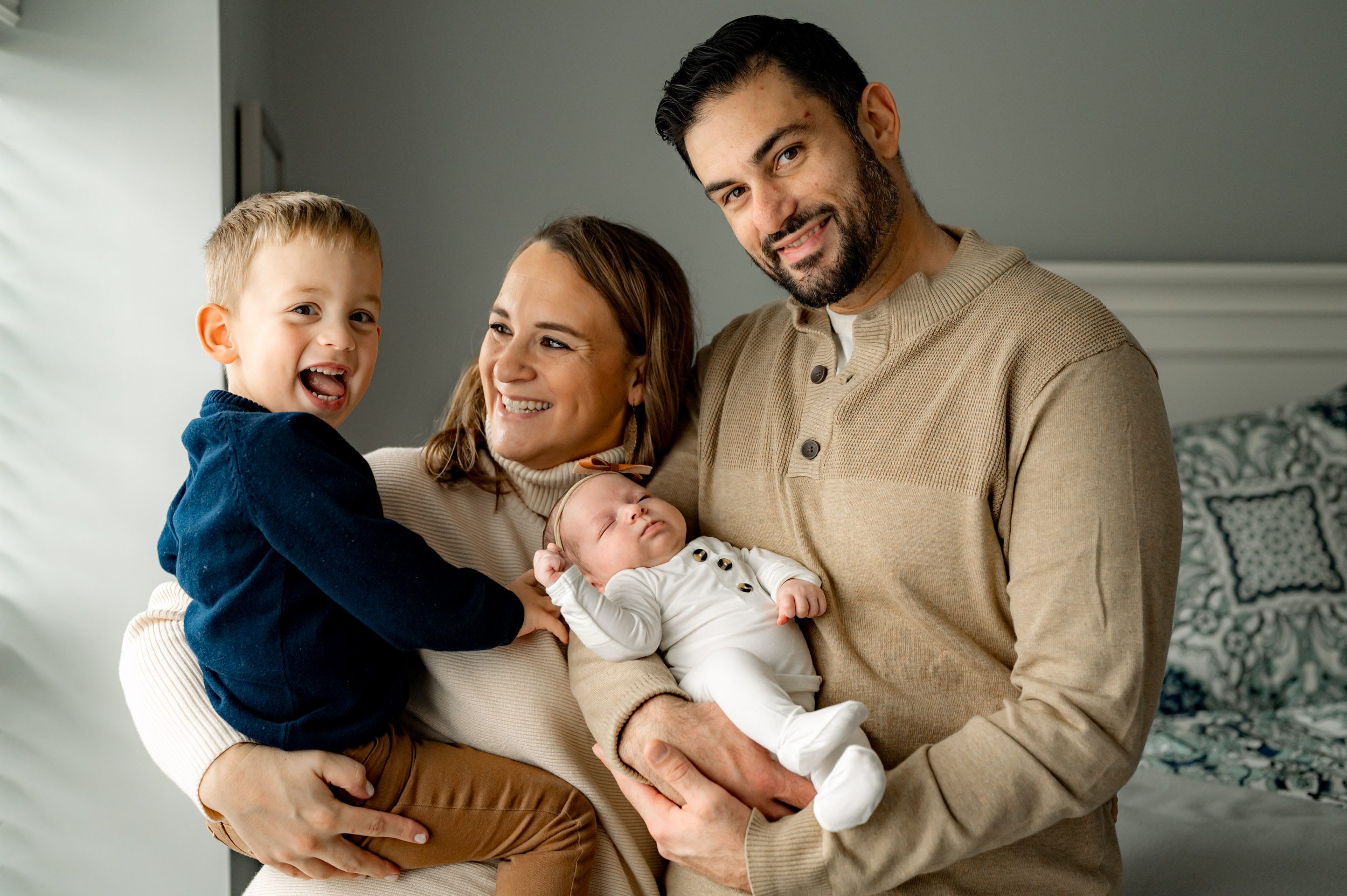 A new family of four holding their baby girl and young son in their arms as big brother smiles and sticks his tongue out at the camera during a newborn photo session