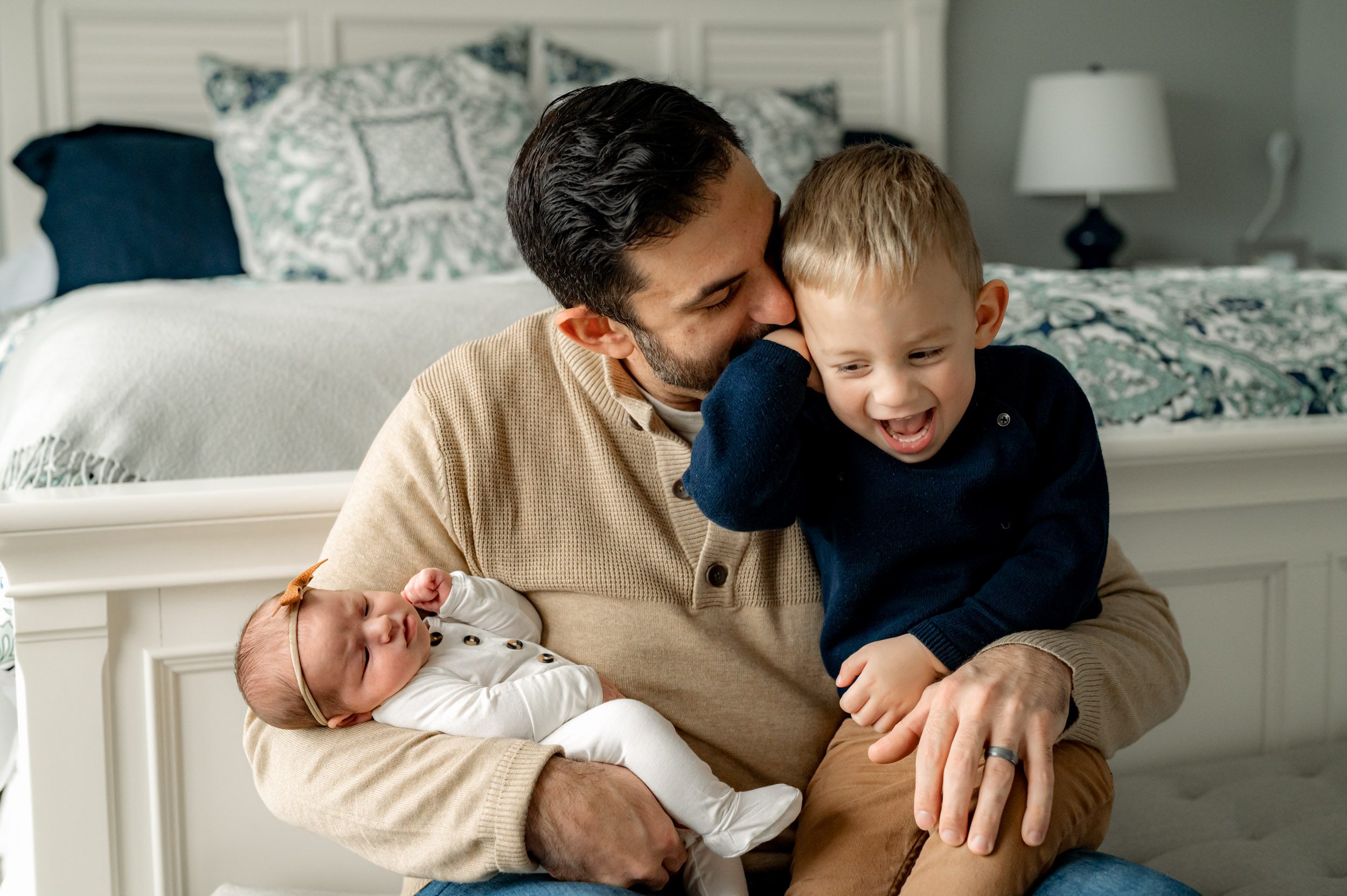 a picture of a dad cradling his baby girl in his arms and kissing his young son on the cheek while his son laughs during a newborn photo session