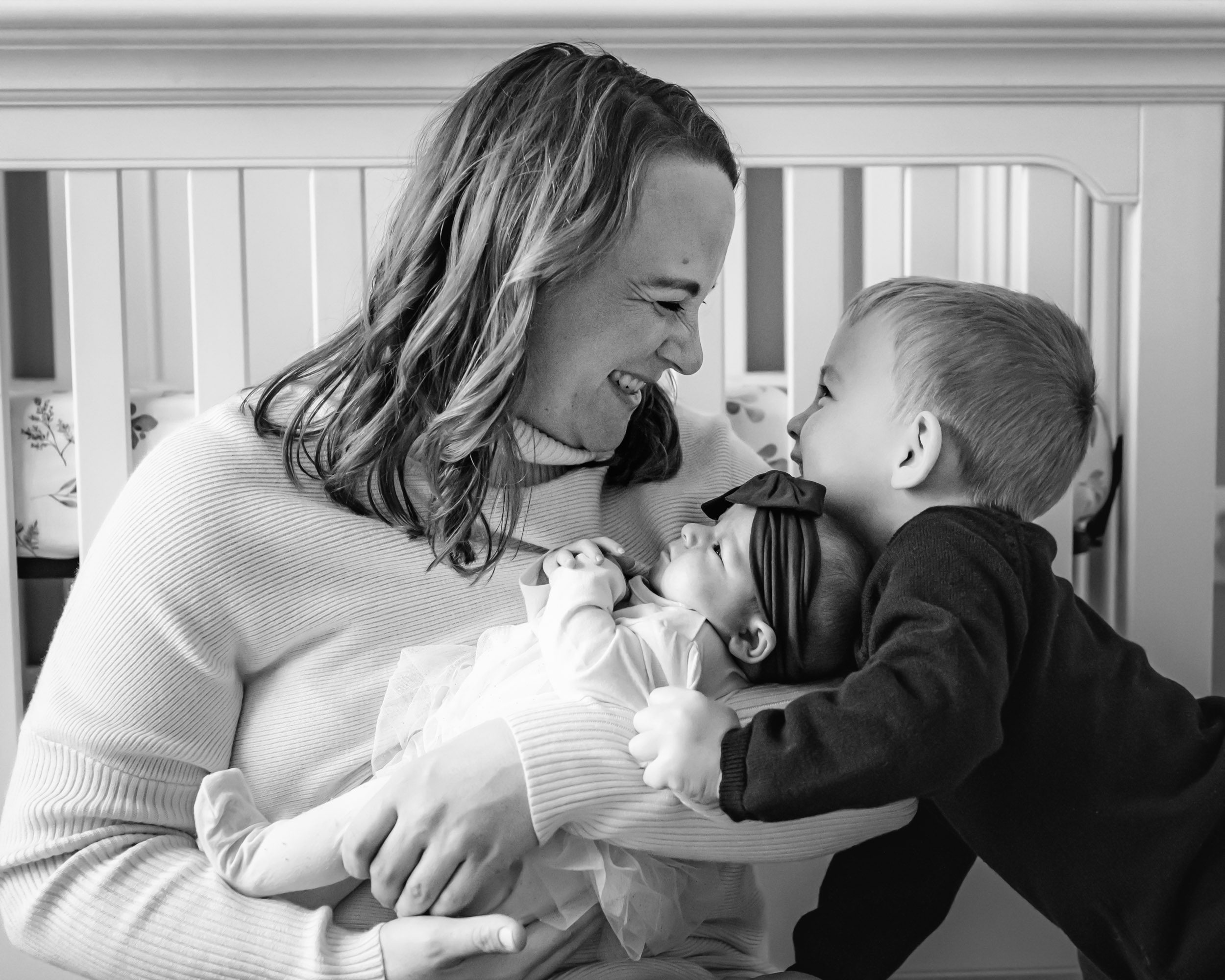 a black and white picture of a mom cradling her baby girl in her arms and smiling at her young son as he hugs his baby sister's head during a newborn photo session