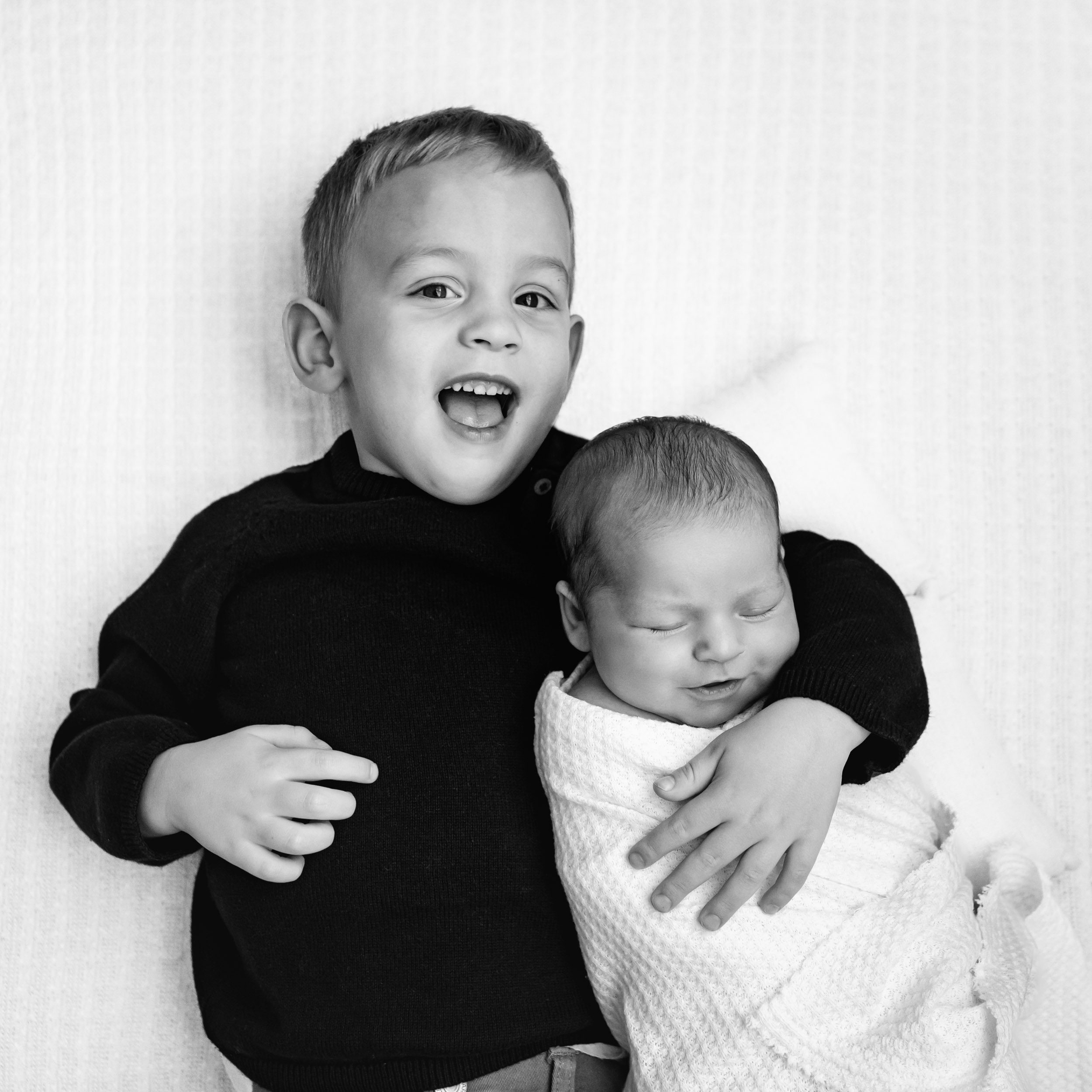 a black and white picture of a young boy laying with his arm around his baby sister and smiling at the camera during a newborn photo session
