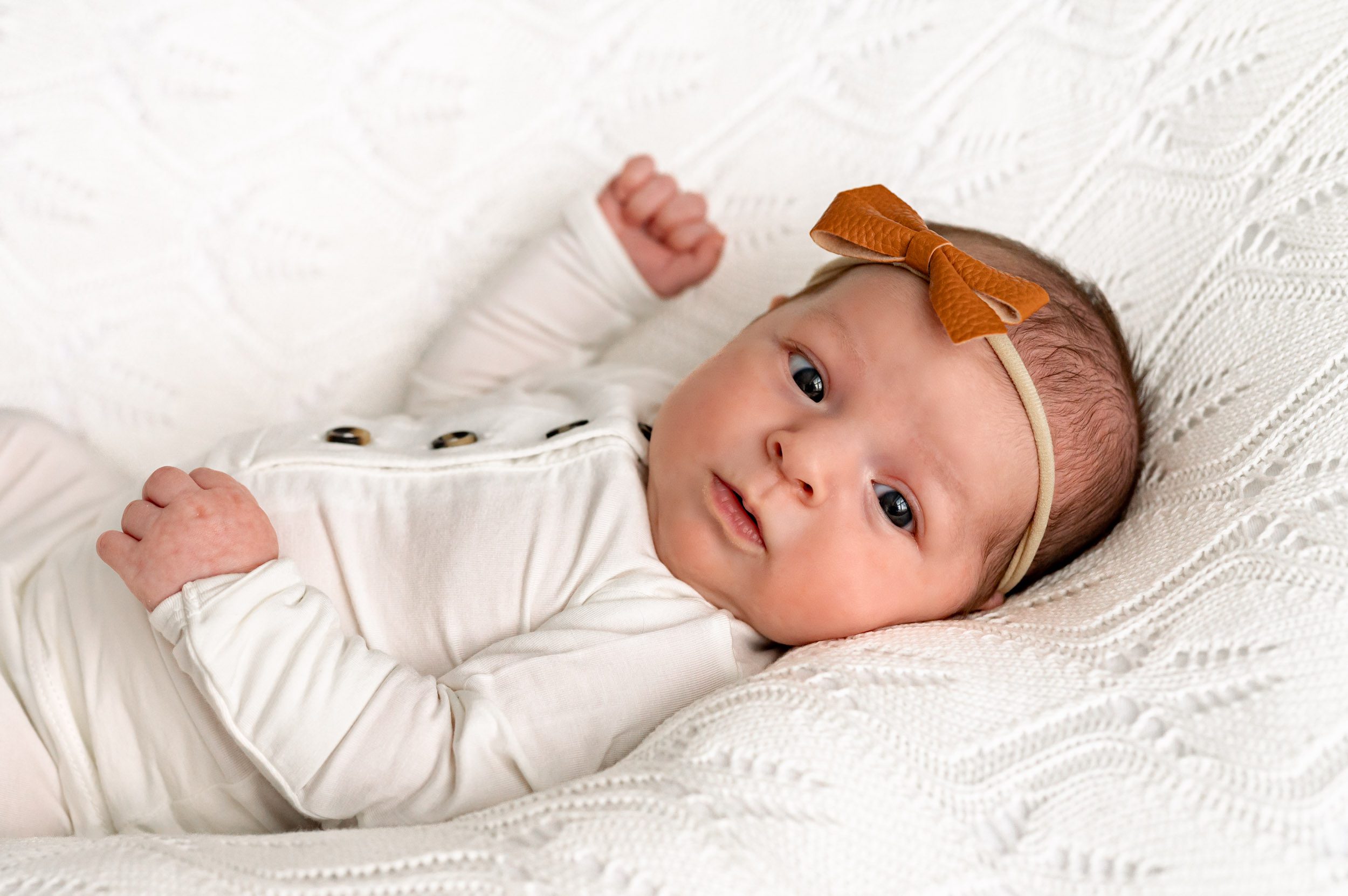 a baby girl in a white body suit laying on a white textured backdrop and looking toward the camera with wide open eyes during a newborn photo session
