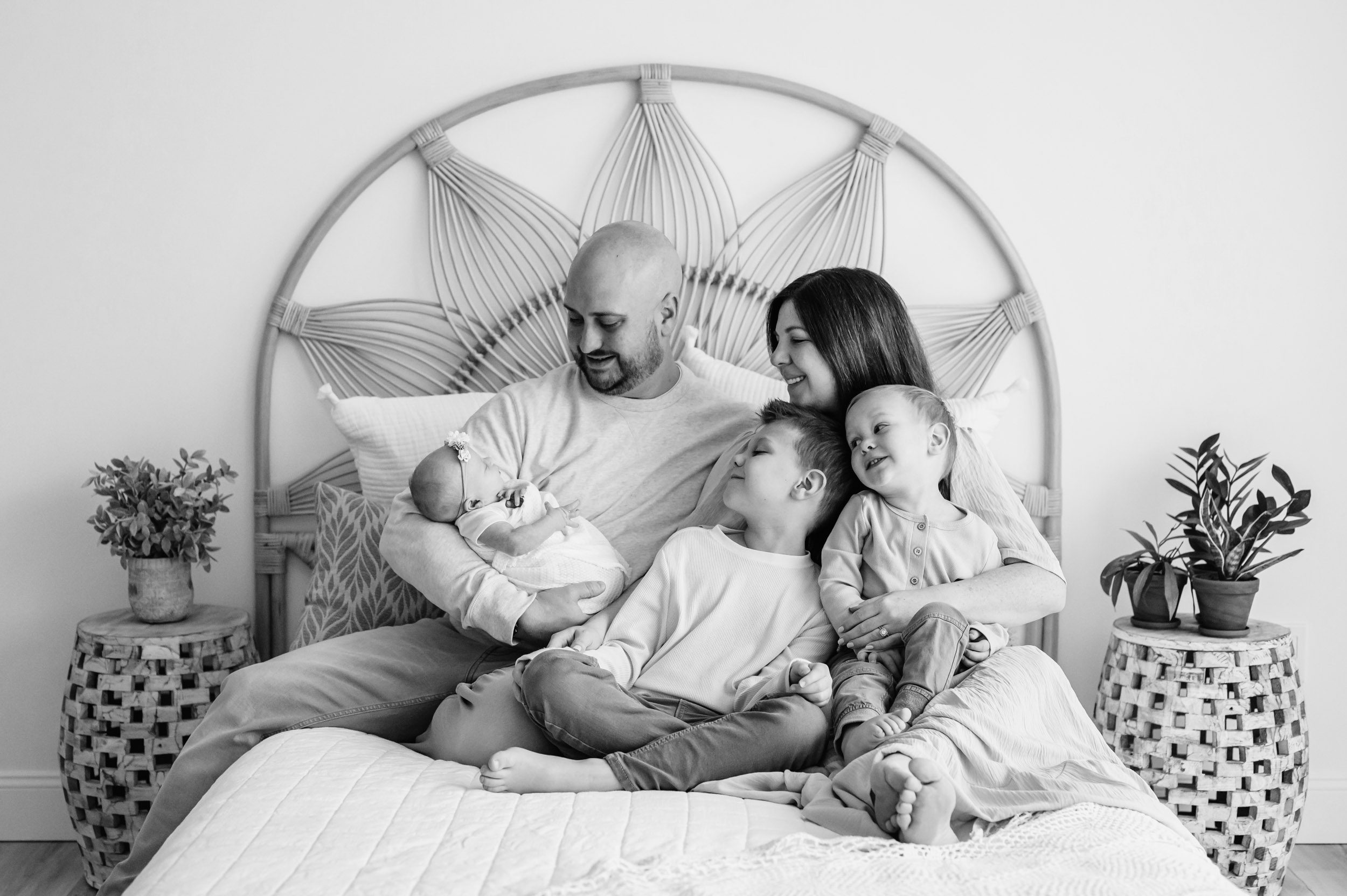 a black and white picture of a family of five sitting on a bed while dad cradles his baby girl in his arms and mom hugs both her sons on her lap as they all look at the baby and smile during a family photoshoot